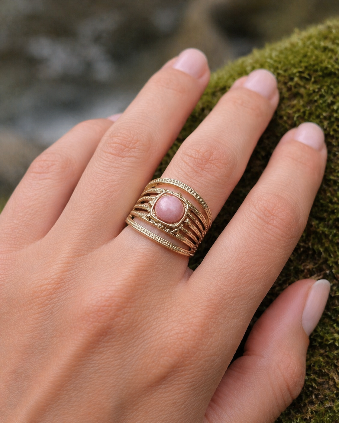 Bague dorée multi-rangs portée au doigt sur décor de mousse, pierre rose ronde et monture travaillée au style bohème élégant.