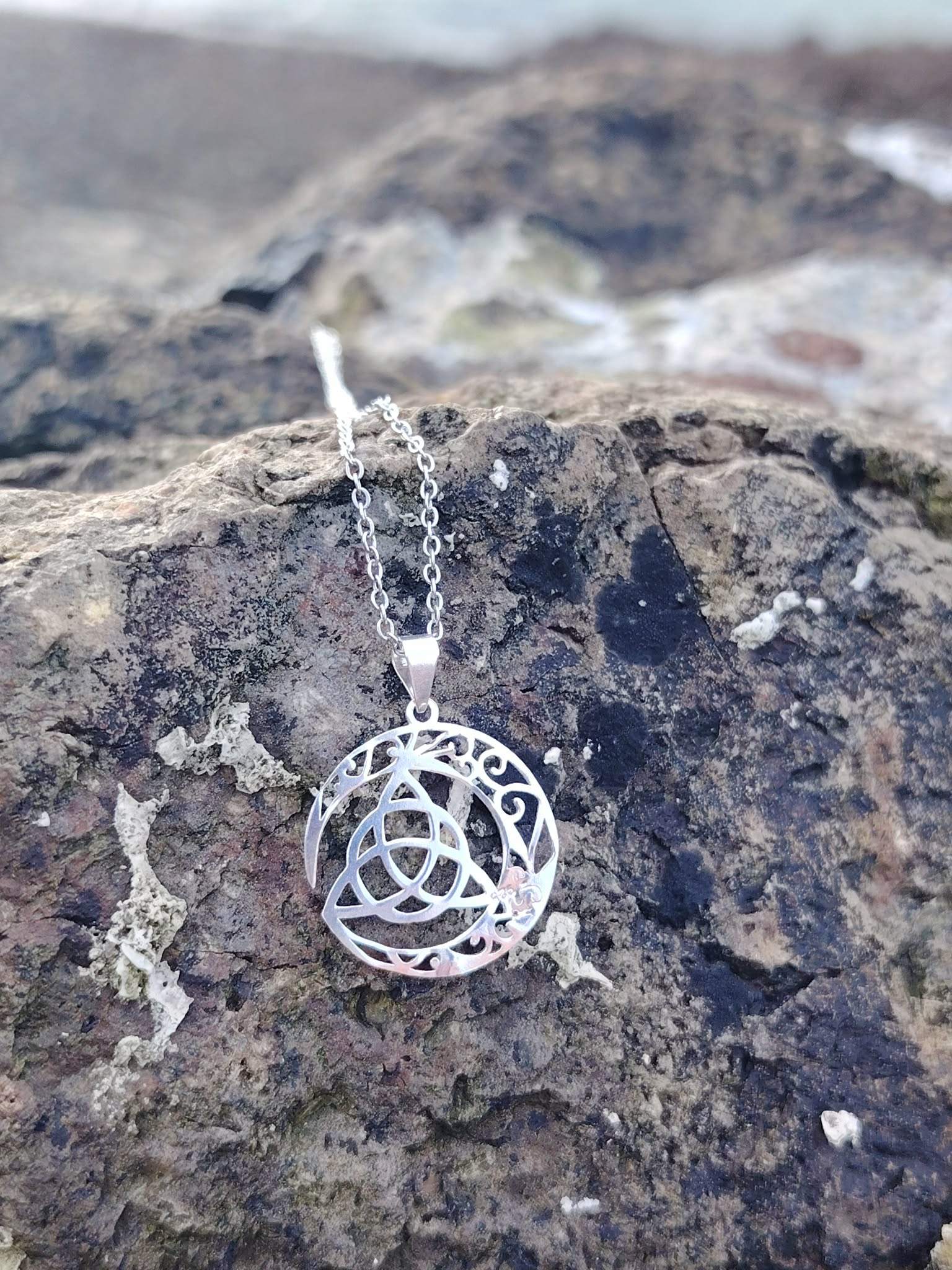 Pendentif Triquetra Lune en argent 925 sur rocher en bord de mer, symbole celtique ajouré et reflets lumineux.