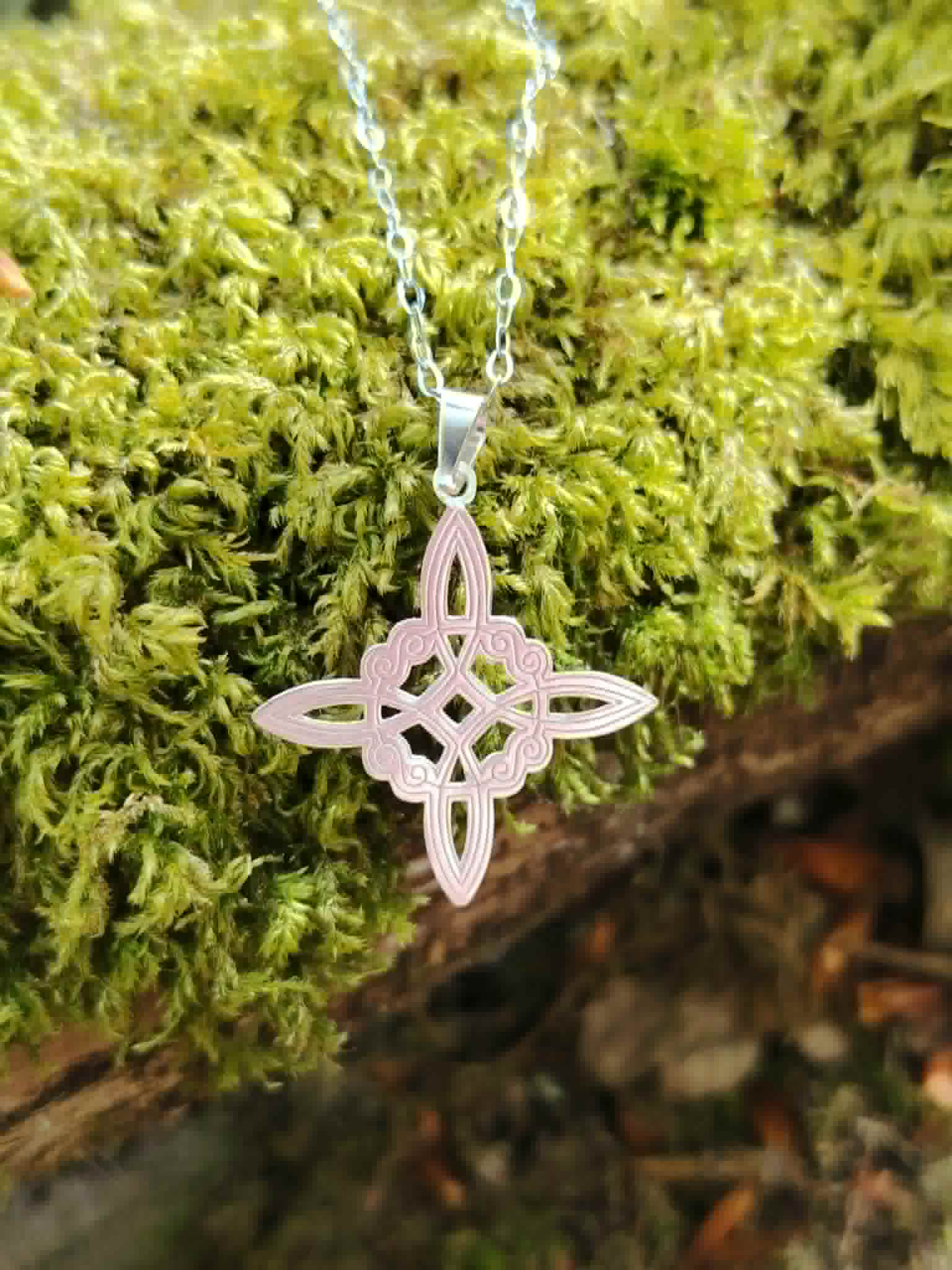 Pendentif Nœud de Sorcière argenté photographié sur mousse bretonne, symbole fin au style ésotérique et naturel.
