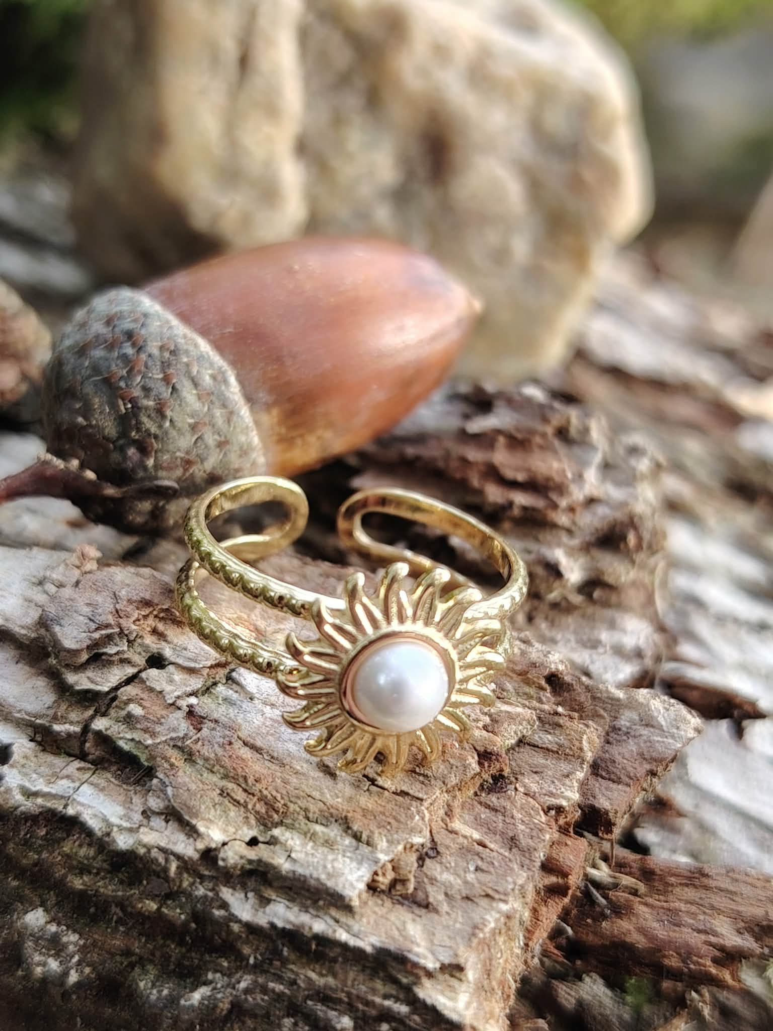 Bague dorée ajustable Soleil avec cabochon blanc nacré posée sur écorce et glands, bijou lumineux en décor naturel breton.
