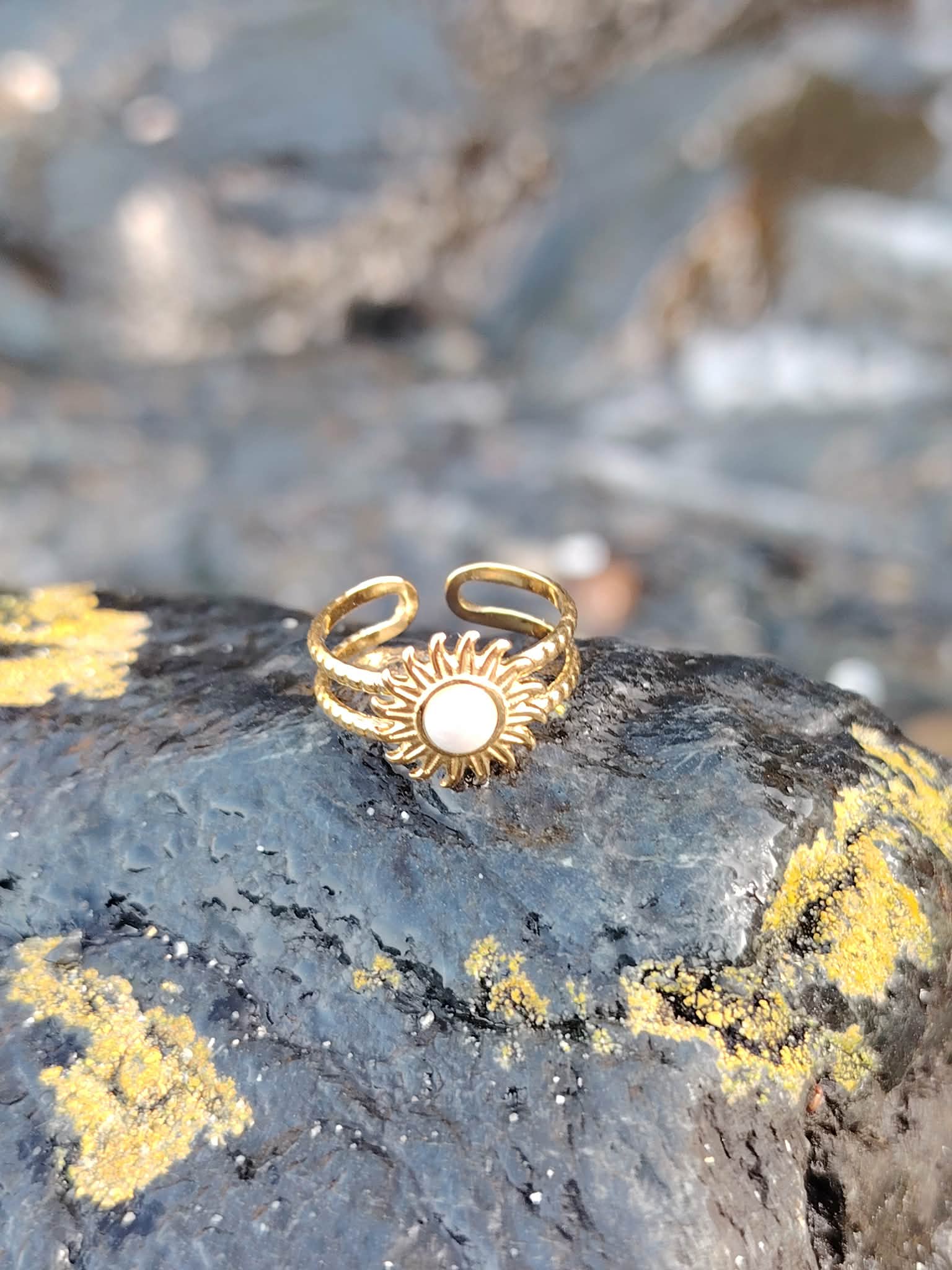 Bague Soleil dorée sur roche sombre avec lichens jaunes, centre nacré et rendu éclatant au bord de mer.