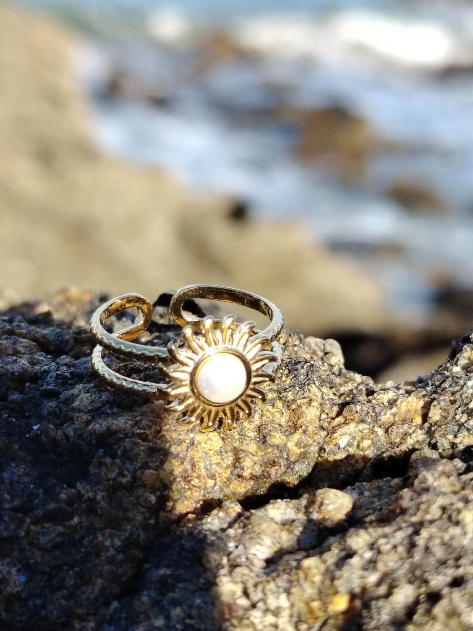 Bague Soleil cabochon nacré photographiée sur roche côtière, bijou doré lumineux avec mer floue en arrière-plan.