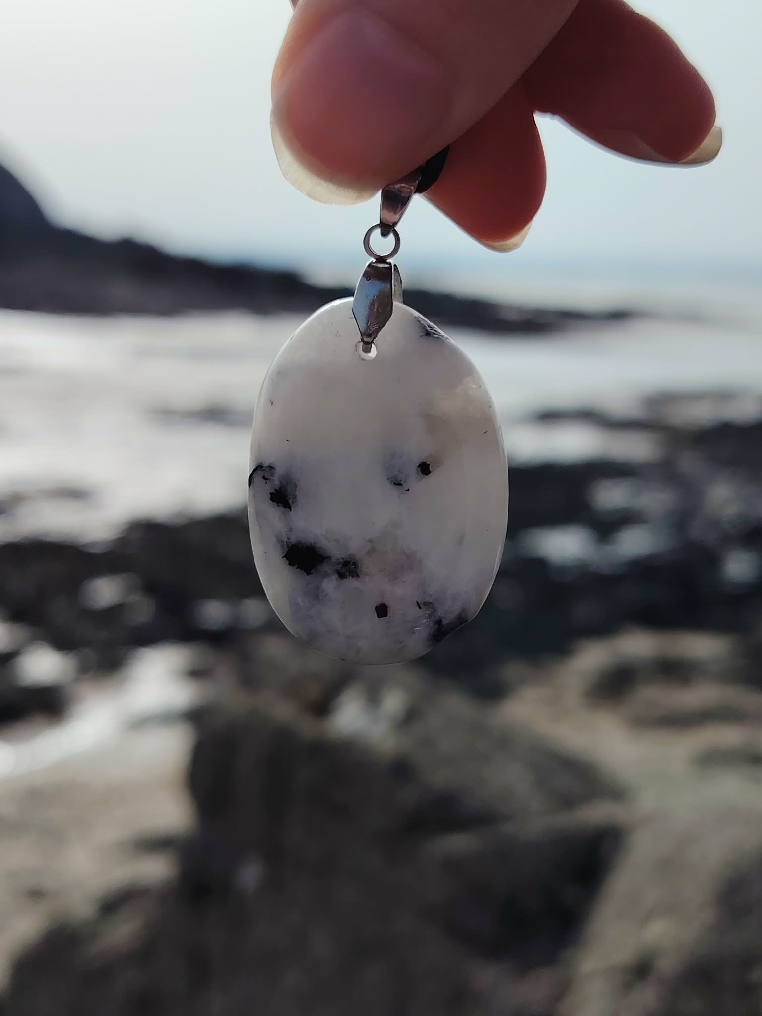 Pendentif Péristérite tenu devant les rochers et la mer en Bretagne, pierre lunaire blanche aux taches naturelles.