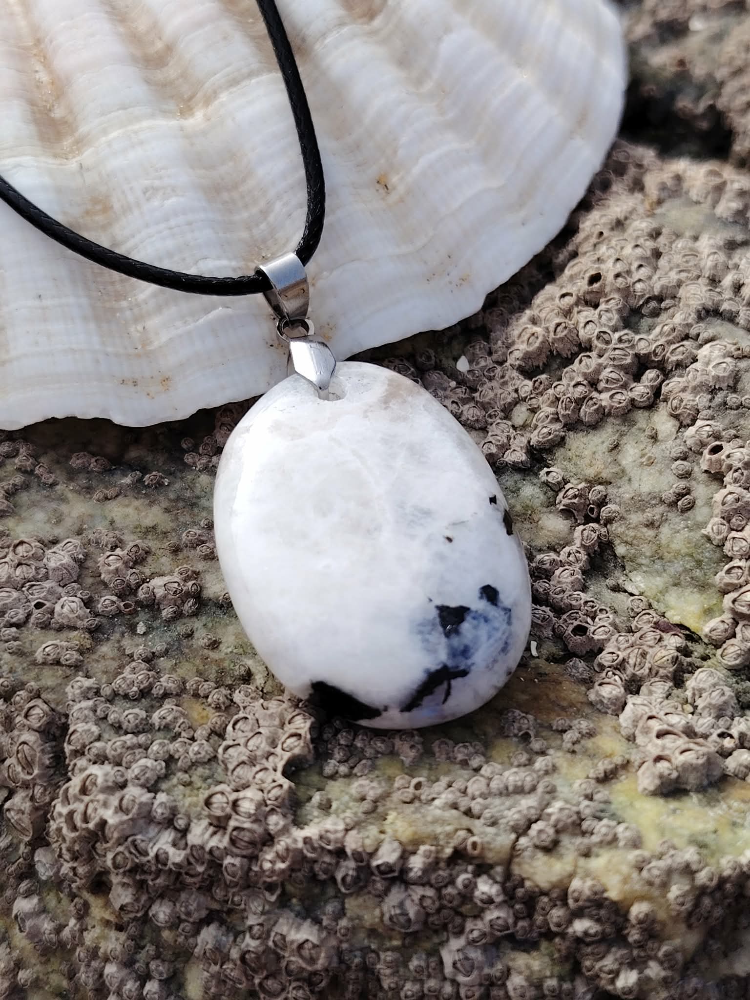 Pendentif Péristérite en gros plan sur décor côtier breton, cordon noir et pierre blanche aux éclats bleutés discrets.