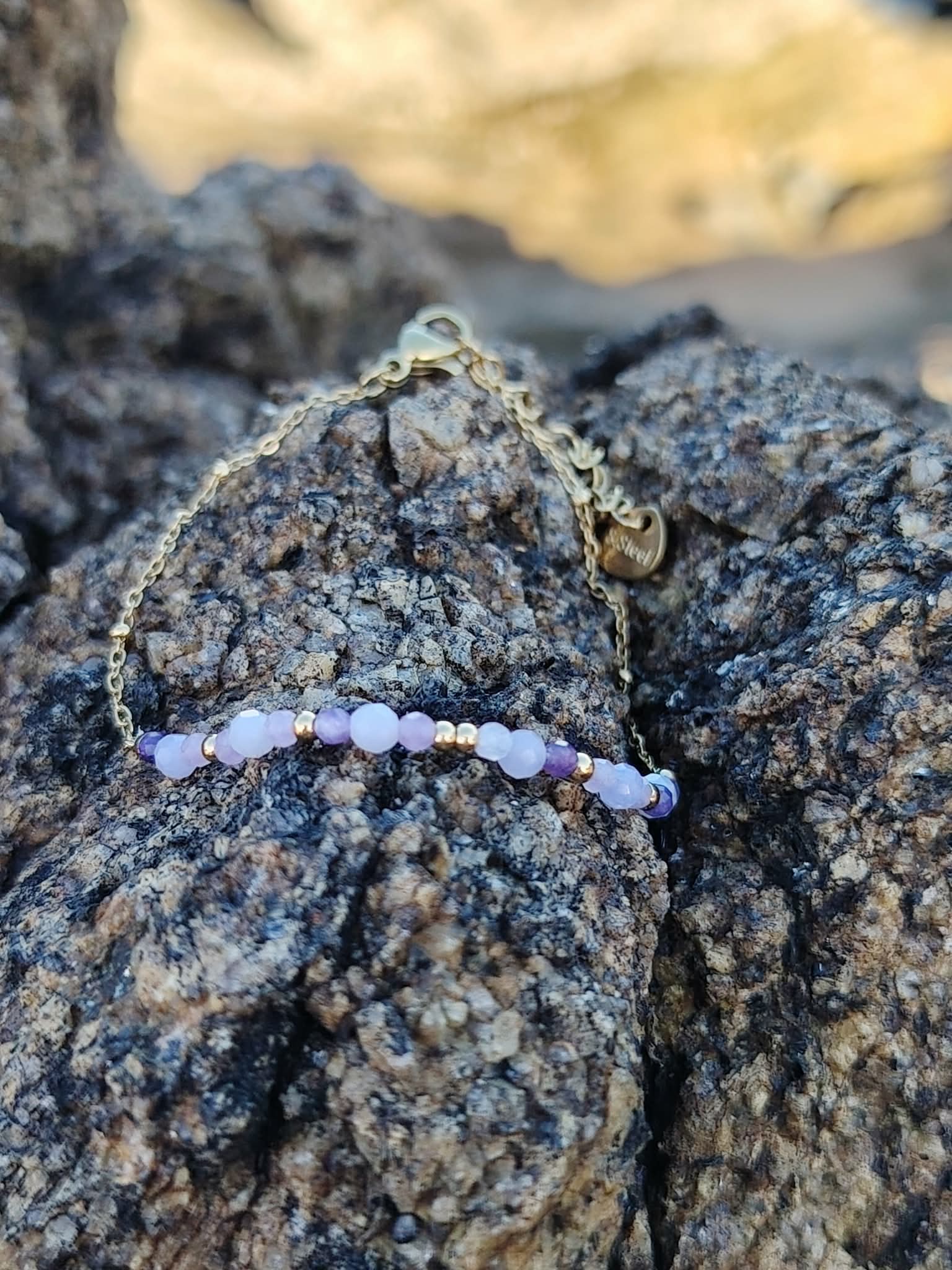 Bracelet Améthyste facettée sur rocher côtier breton, chaîne fine dorée et breloques pour équilibre émotionnel.