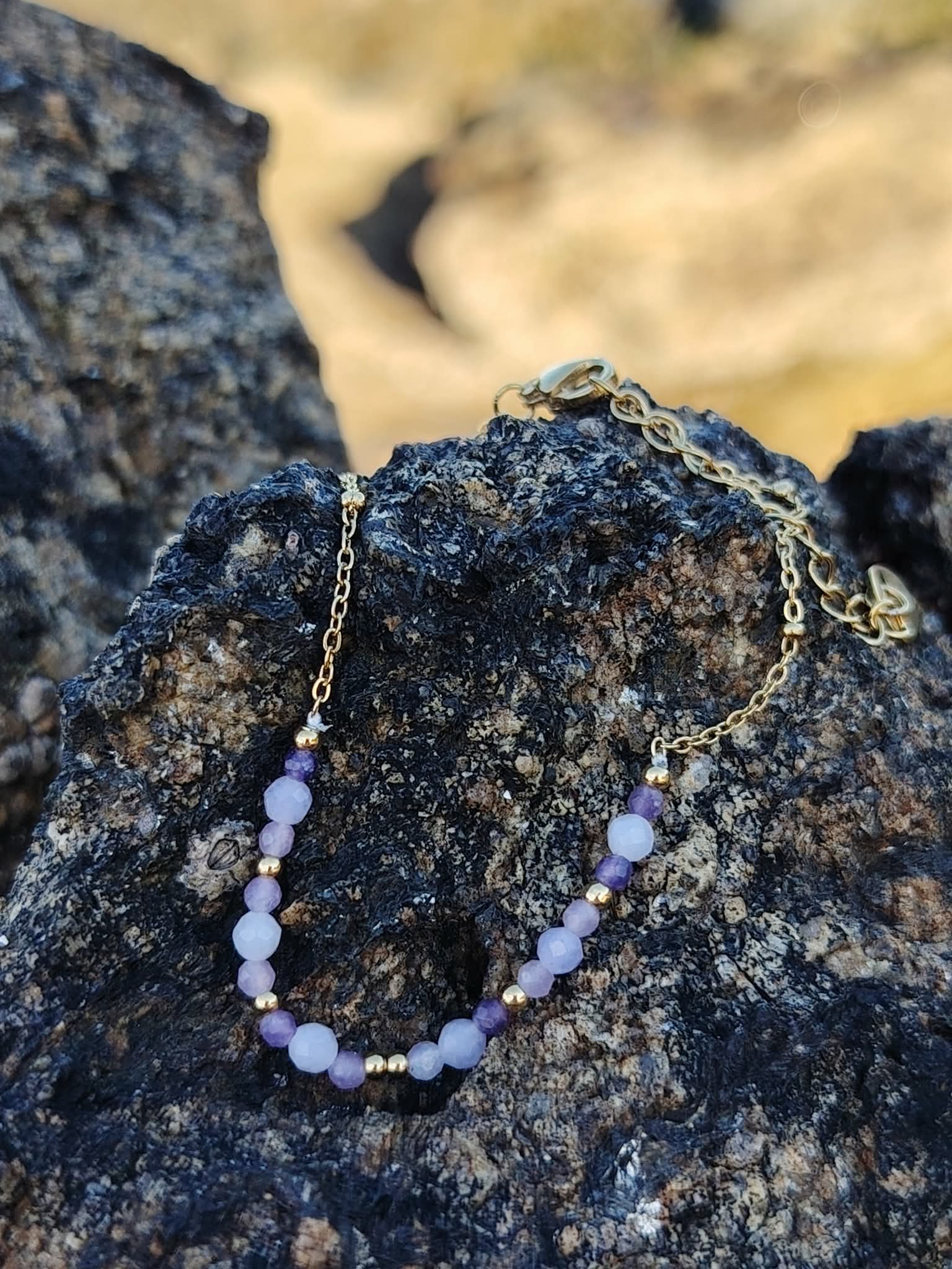 Bracelet Améthyste facettée doré posé sur roche en bord de mer breton, perles violettes et breloques pour sérénité.