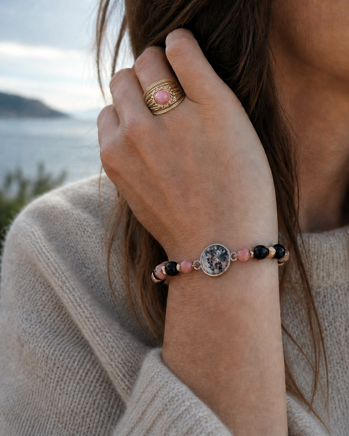 Bracelet rose, noir et doré porté au poignet avec bague assortie, face à la mer en Bretagne, élégance douce et harmonie émotionnelle.