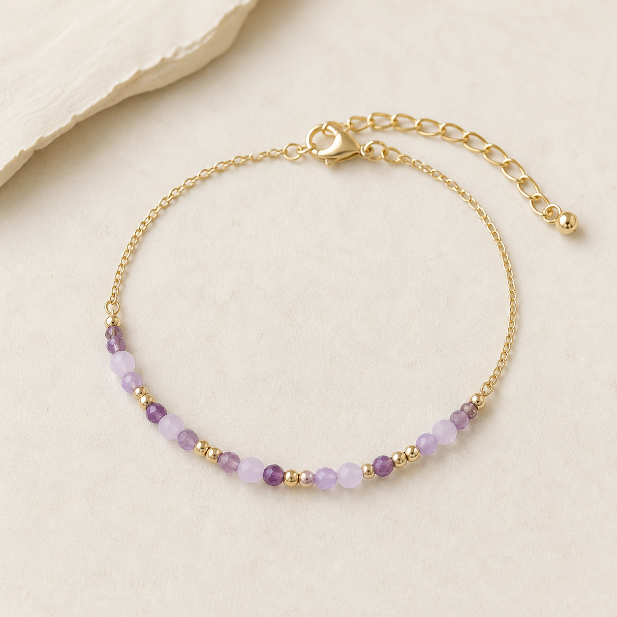 Bracelet améthyste facetté 3 mm sur chaîne dorée, perles lilas et parme avec petits intercalaires dorés sur fond beige élégant.
