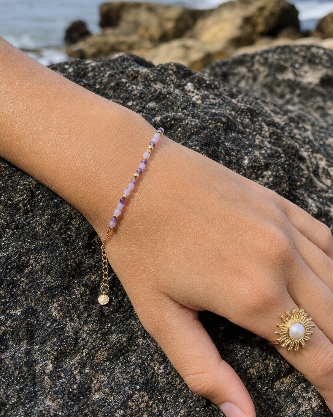 Main posée sur un rocher en bord de mer, bracelet fin en améthyste facettée sur chaîne dorée avec breloque et bague soleil dorée à perle blanche, dans une ambiance lumineuse évoquant sérénité et intuition.
