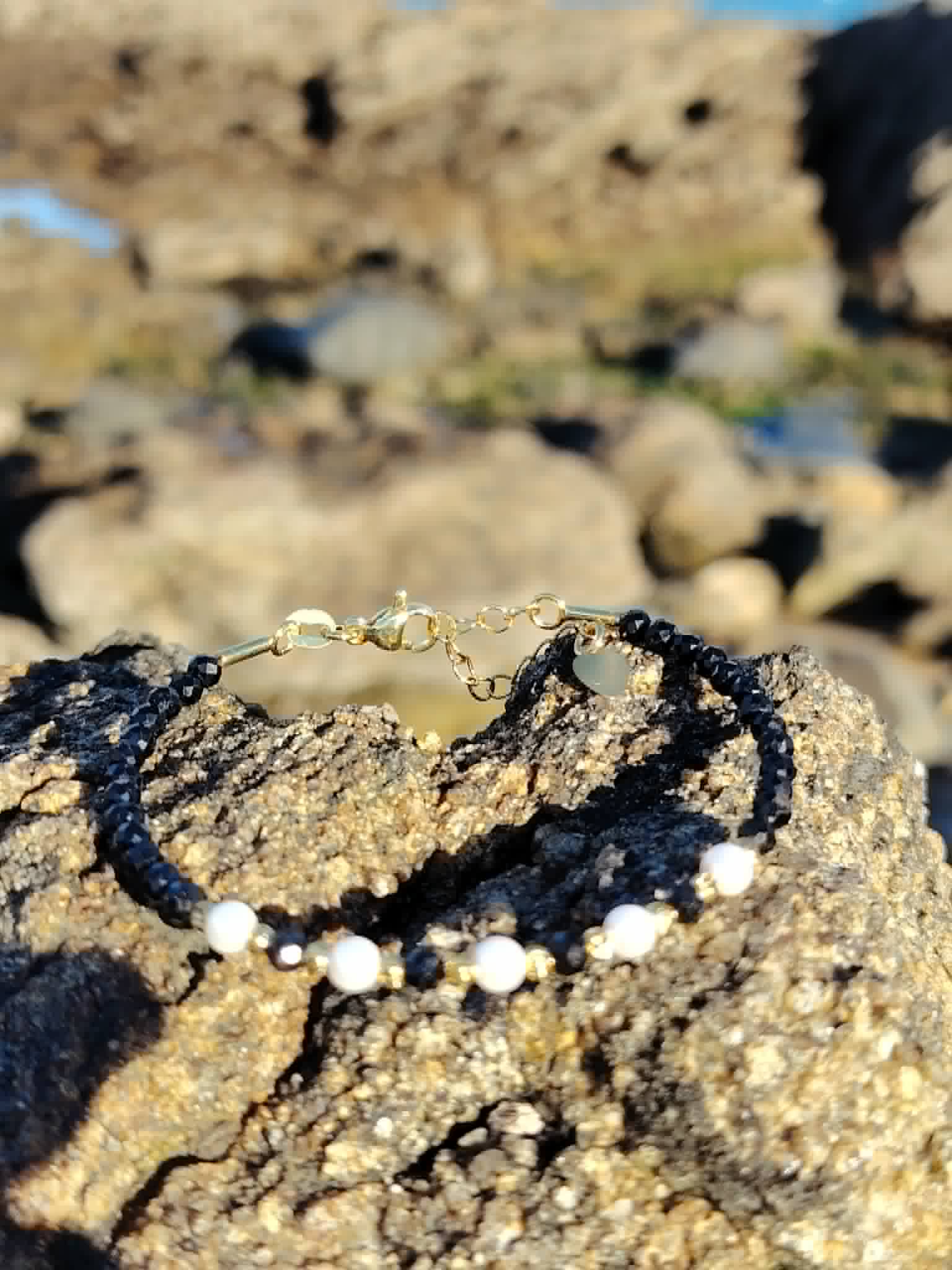 Bracelet fin en tourmaline noire facettée avec perles blanches et détails dorés, photographié sur rocher en lumière naturelle, style délicat et protecteur.