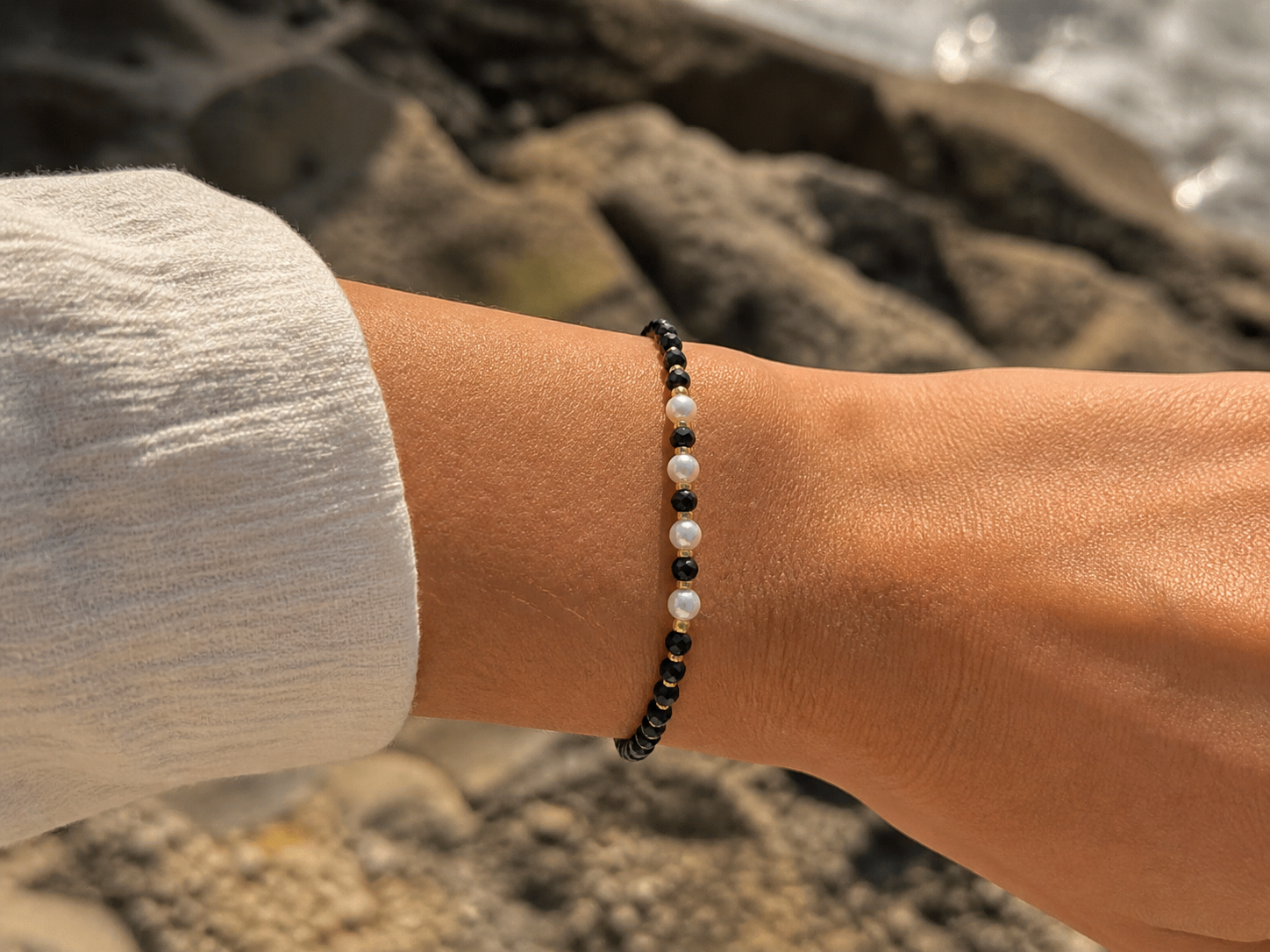 Bracelet Tourmaline Noire facettée 2 mm porté au poignet près de la mer sur rochers, perles noires, blanches et dorées pour protection et élégance discrète.