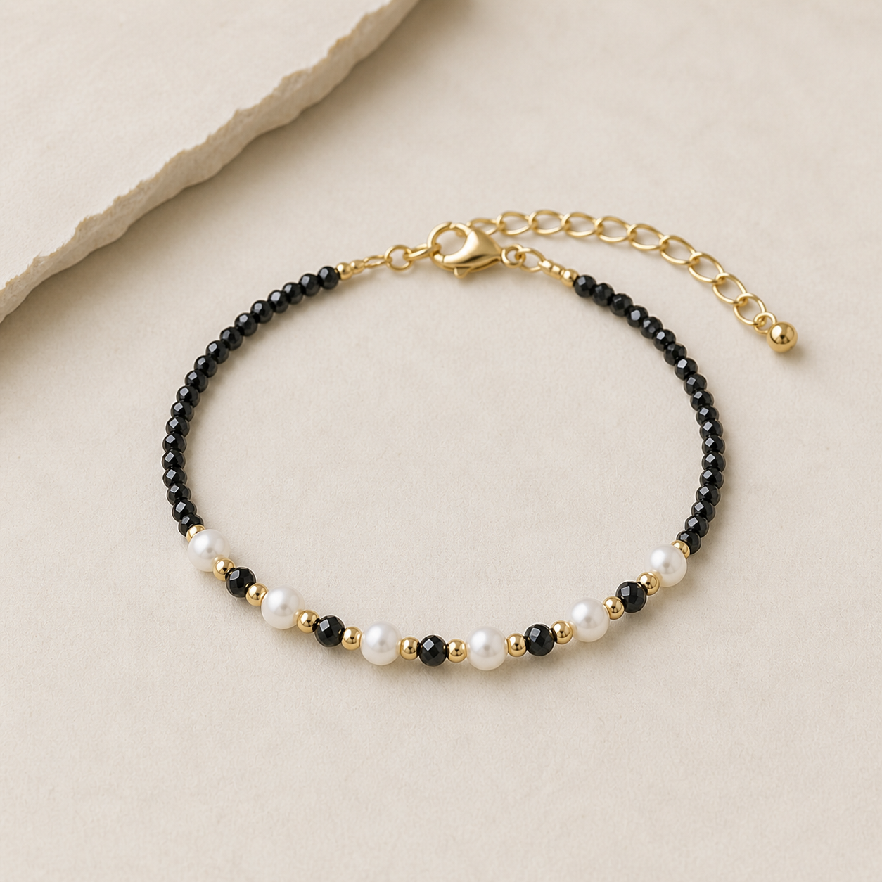 Bracelet fin en Tourmaline Noire facettée 2 mm avec perles blanches et détails dorés sur fond beige, bijou ajustable pour ancrage et style raffiné.