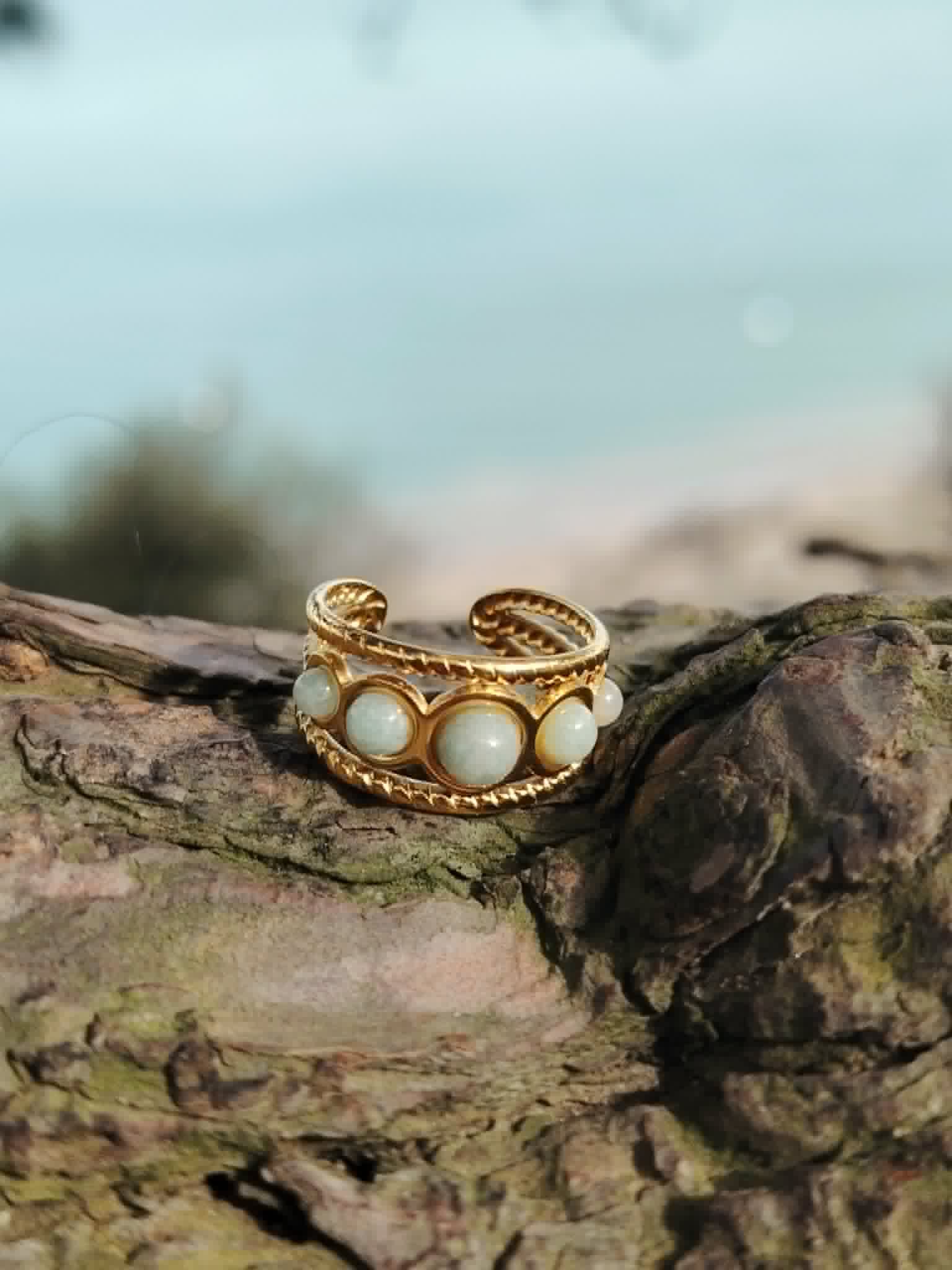 Bague dorée réglable multi Amazonite posée sur bois avec arrière-plan marin flou, cabochons bleu-vert pour apaisement et élégance.