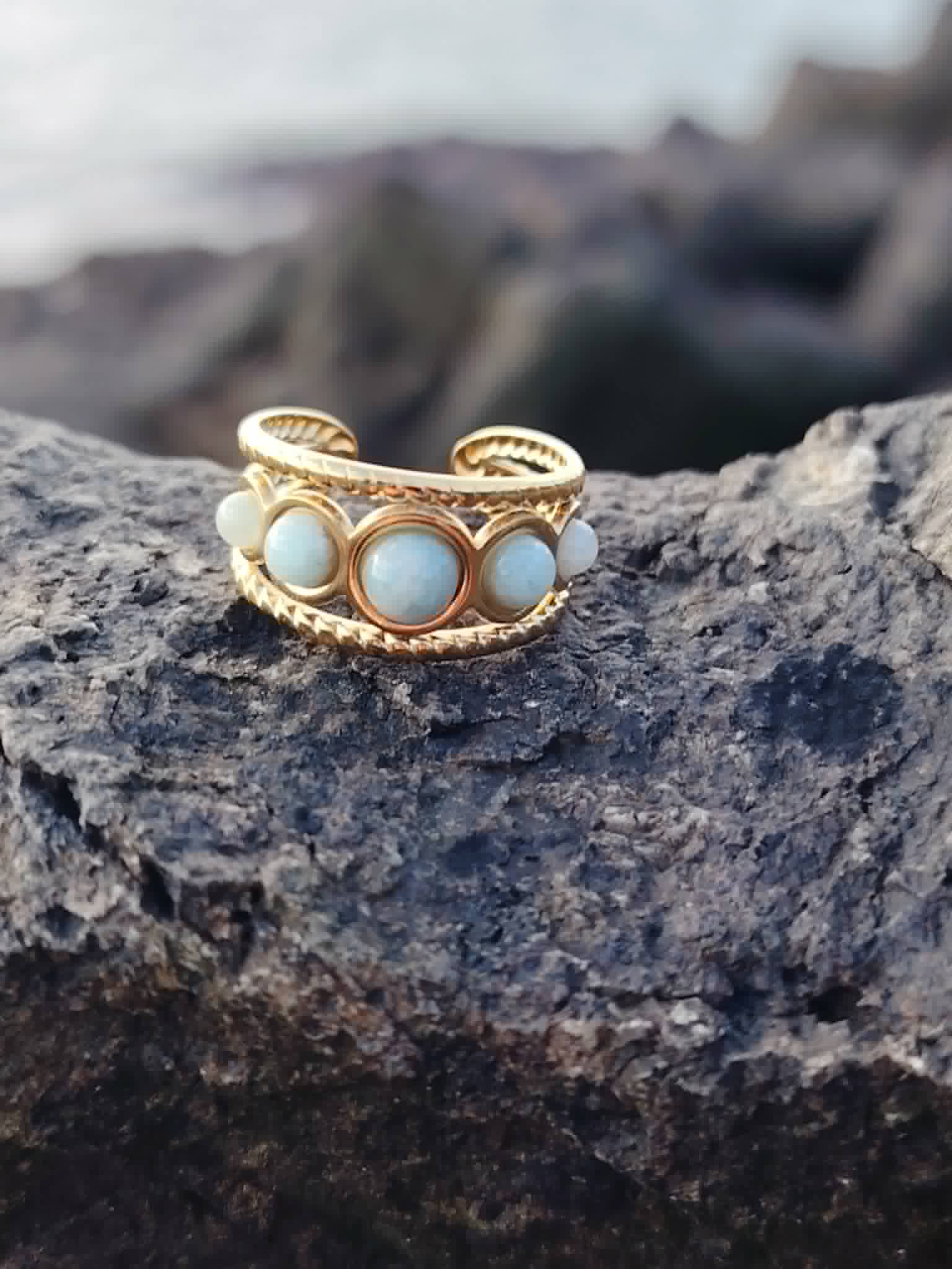 Bague dorée multi Amazonite posée sur roche près de la mer, cabochons lumineux et monture ajourée pour calme et style solaire.