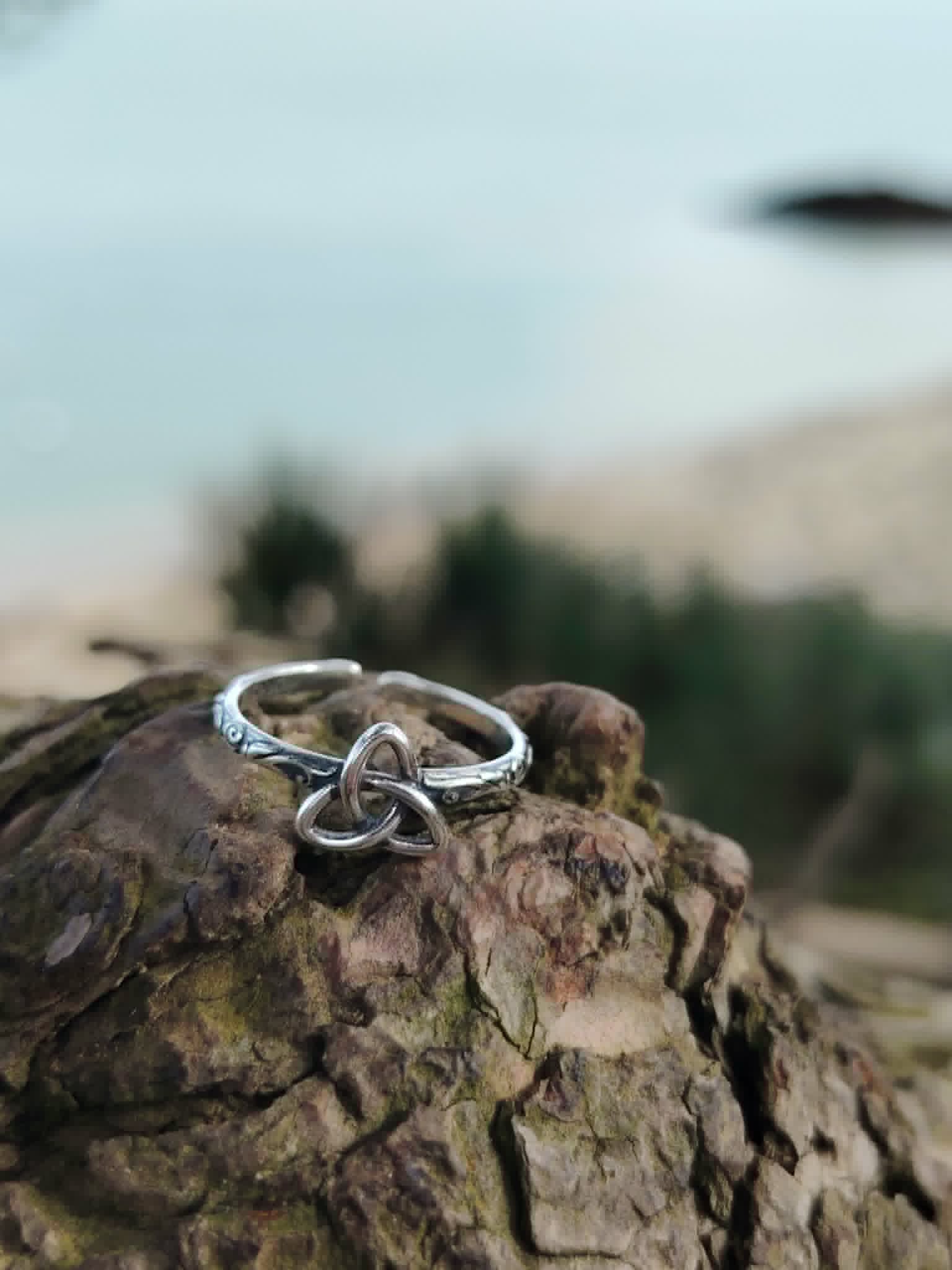Bague argentée Triquetra posée sur tronc avec mer floue en arrière-plan, bijou ajustable évoquant unité, cycles et équilibre intérieur.