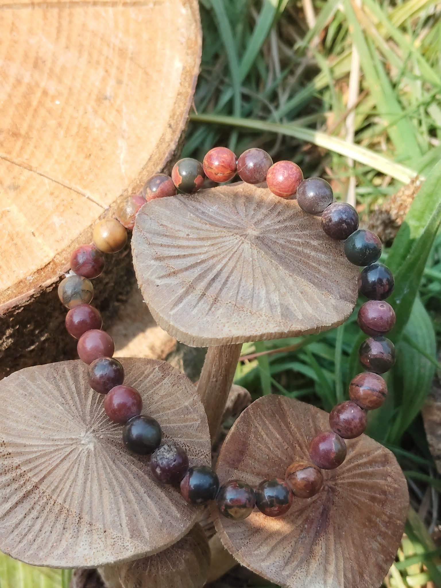 Bracelet Jaspe Polychrome présenté sur des champignons décoratifs en bois avec herbes autour, perles rouge brun et noires, pour retour à soi et équilibre.