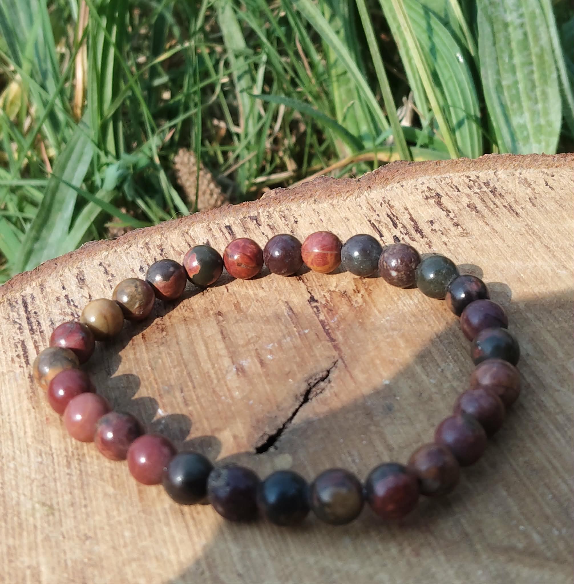 Bracelet Jaspe Polychrome photographié sur une tranche de bois avec herbes en arrière-plan, perles rouges, brunes et noires, pour protection et ancrage profond.
