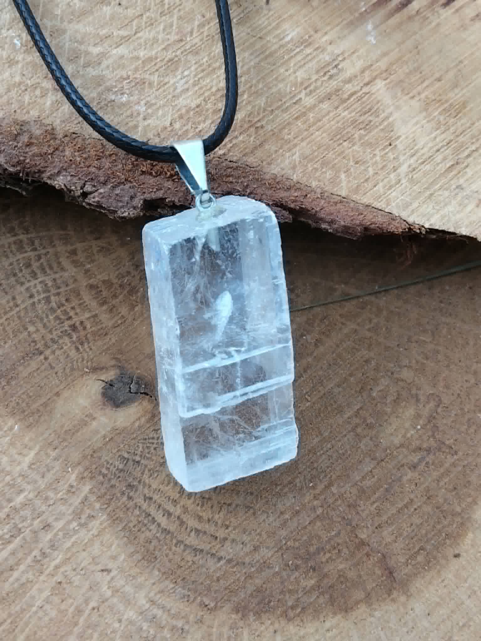 Pendentif Calcite Optique sur cordon noir posé sur bois brut, pierre transparente rectangulaire avec bélière argentée pour intuition et recul intérieur.