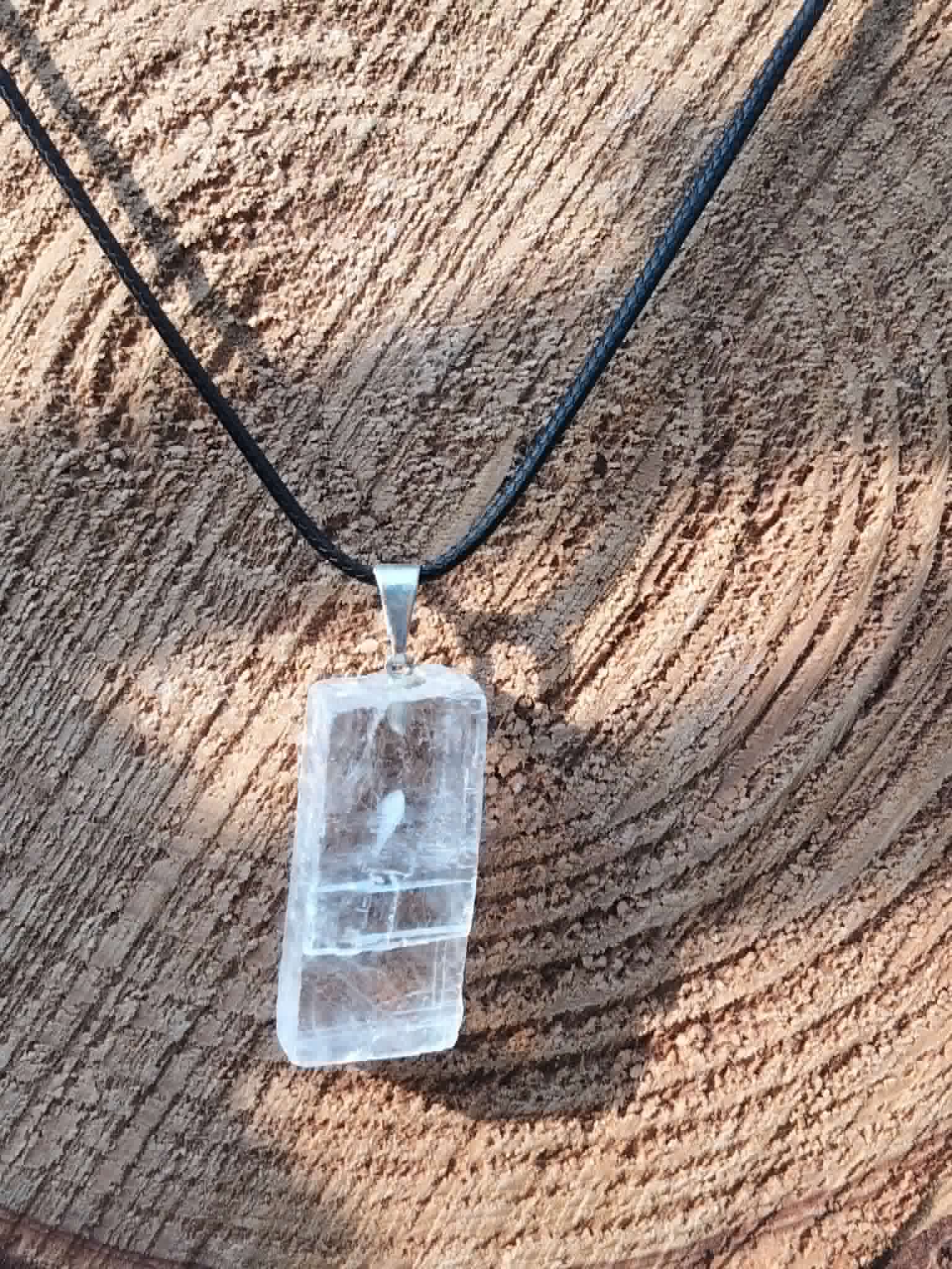 Pendentif Calcite Optique rectangulaire sur bois ensoleillé, cordon noir et reflets givrés pour accompagner les intentions positives.