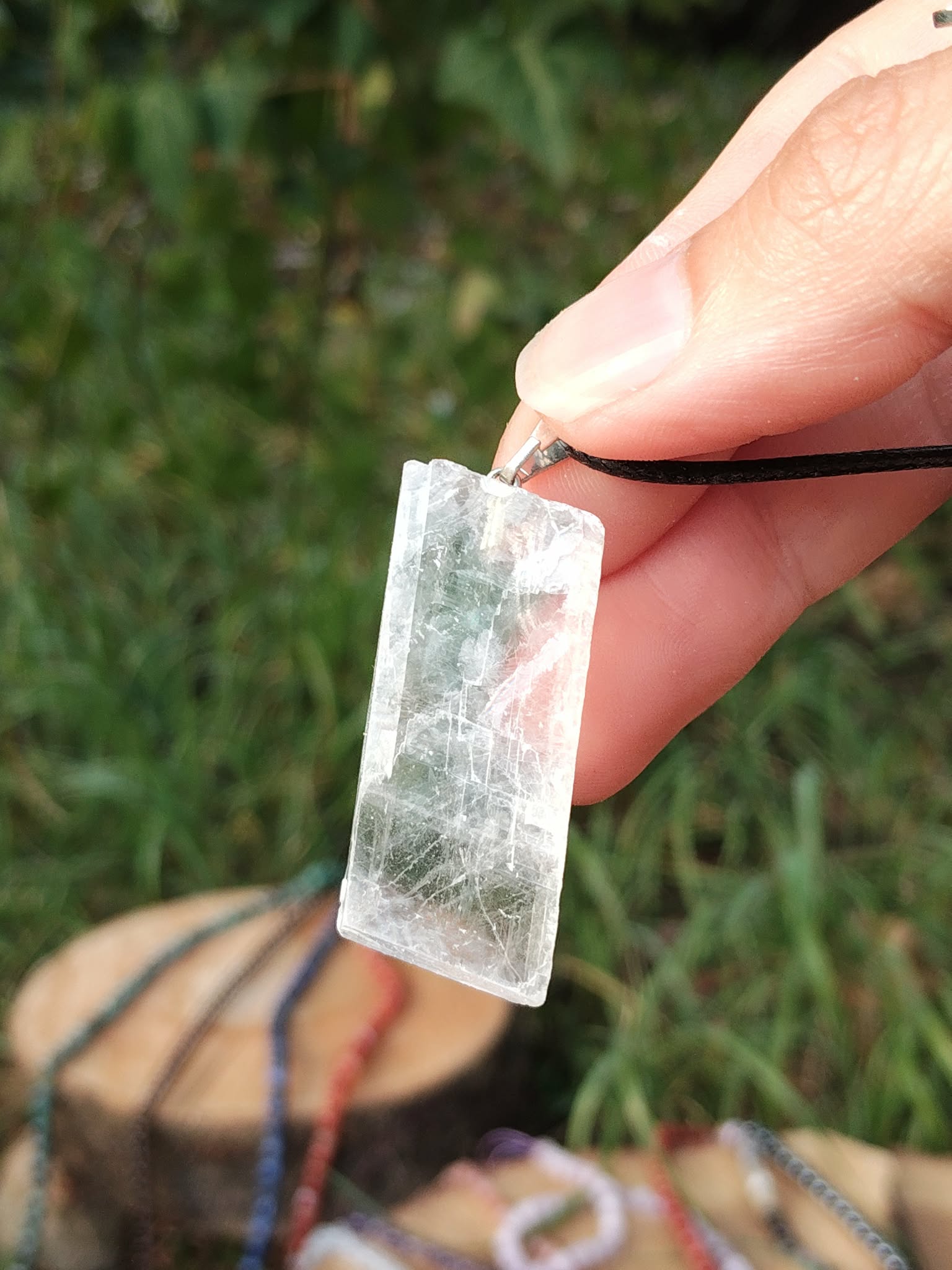 Pendentif Calcite Optique tenu à la main en extérieur, pierre transparente sur cordon noir avec fond végétal pour clarté et intuition.
