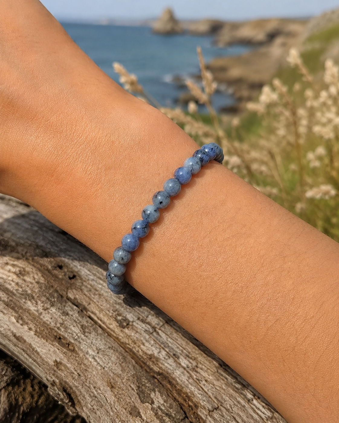 Bracelet Tanzanite perles 6-7 mm porté au poignet sur bois flotté, mer et falaises de Bretagne en arrière-plan, intuition, expression et accomplissement.