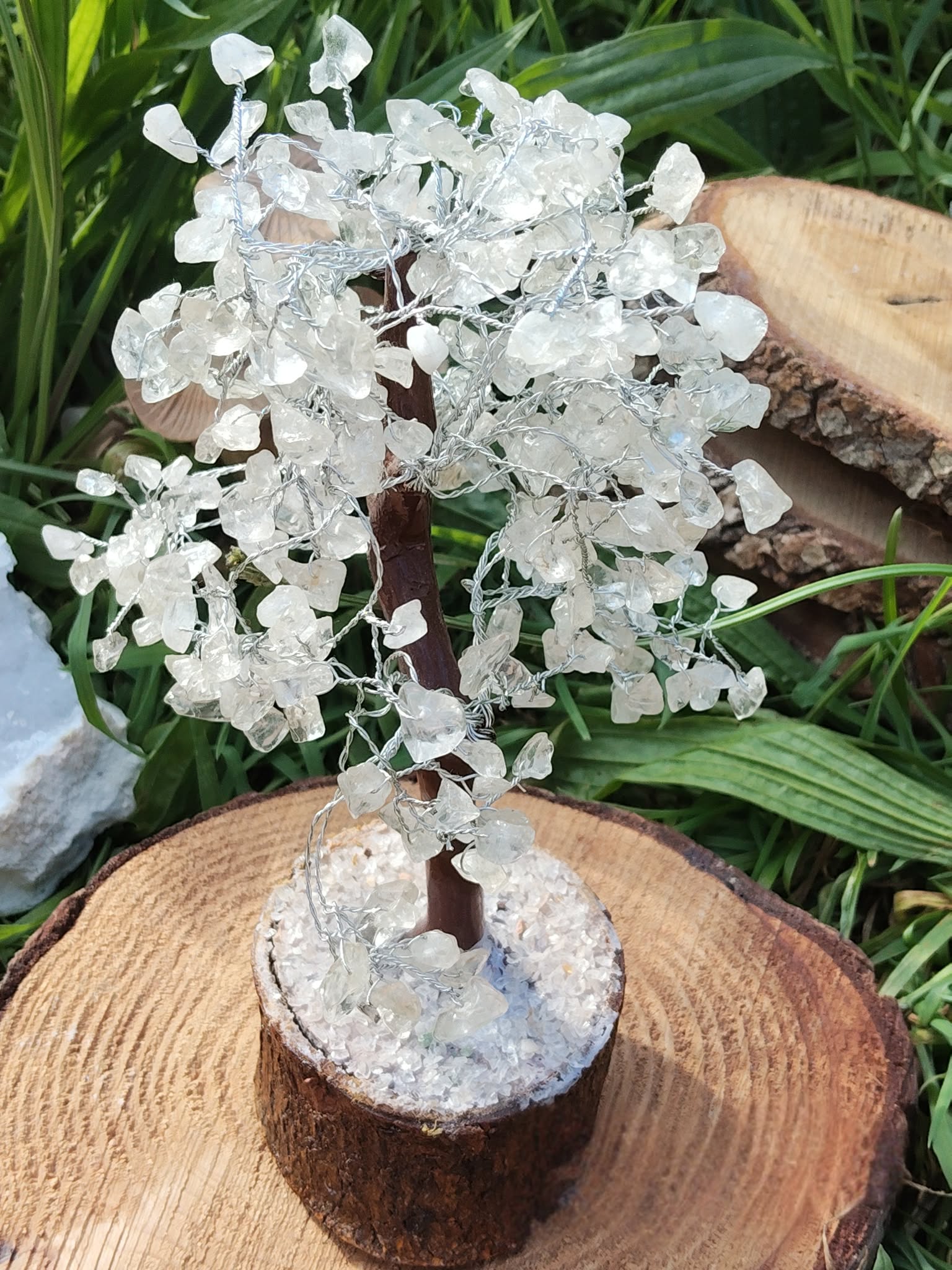 Arbre de vie en cristal de roche sur socle en bois, photographié dans l’herbe avec rondins, clarté et énergie douce.