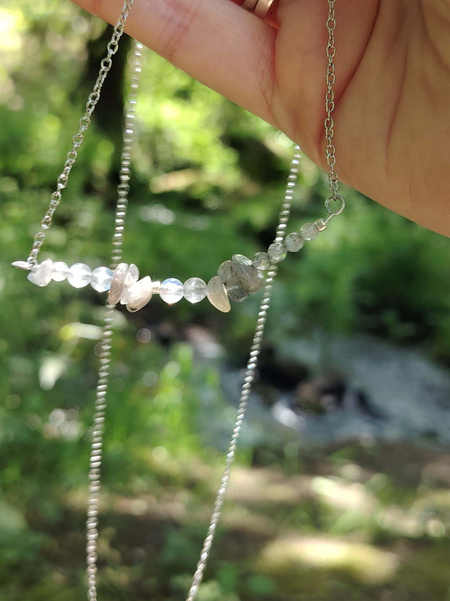 Détail du collier Labradorite tenu à la main, perles claires et reflets froids dans une ambiance boisée.