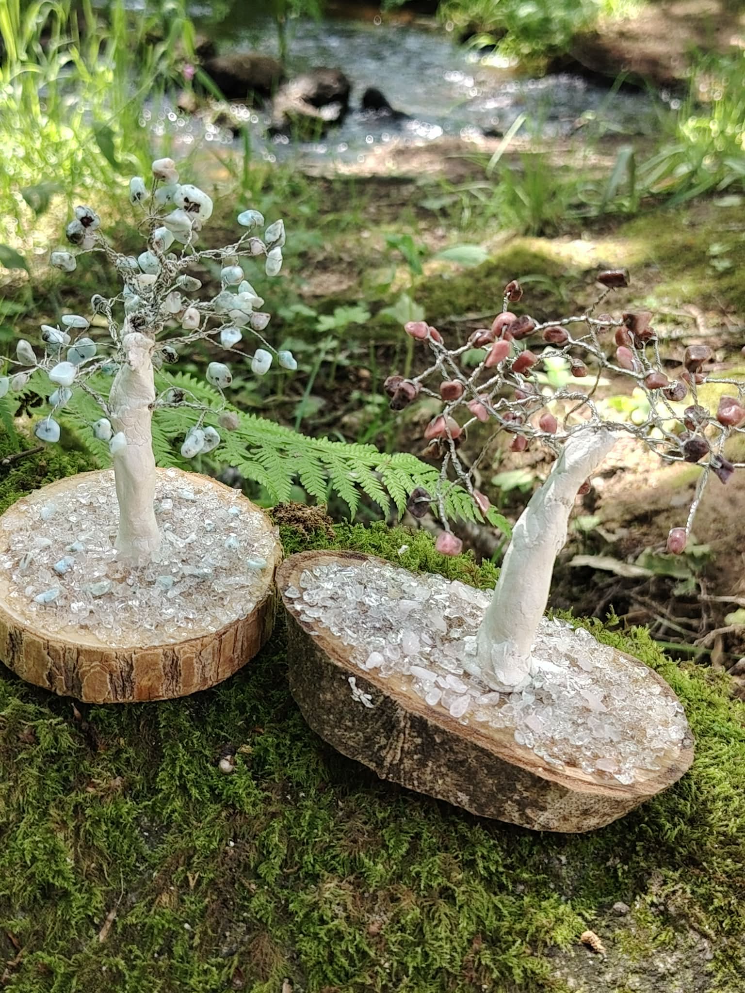 Duo d’arbres Magic Gaïa en rhodonite et pierres bleu pâle sur rondins de bois, décor forestier avec fougères et ruisseau.