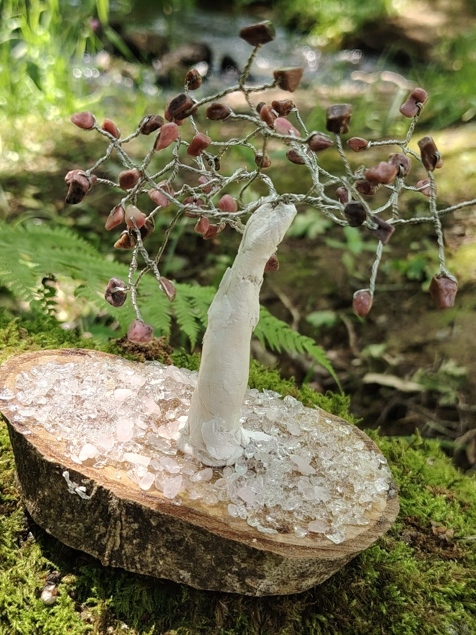 Création Magic Gaïa arbre en rhodonite sur socle bois et cristal, posé sur mousse près d’un ruisseau en forêt, douceur émotionnelle.