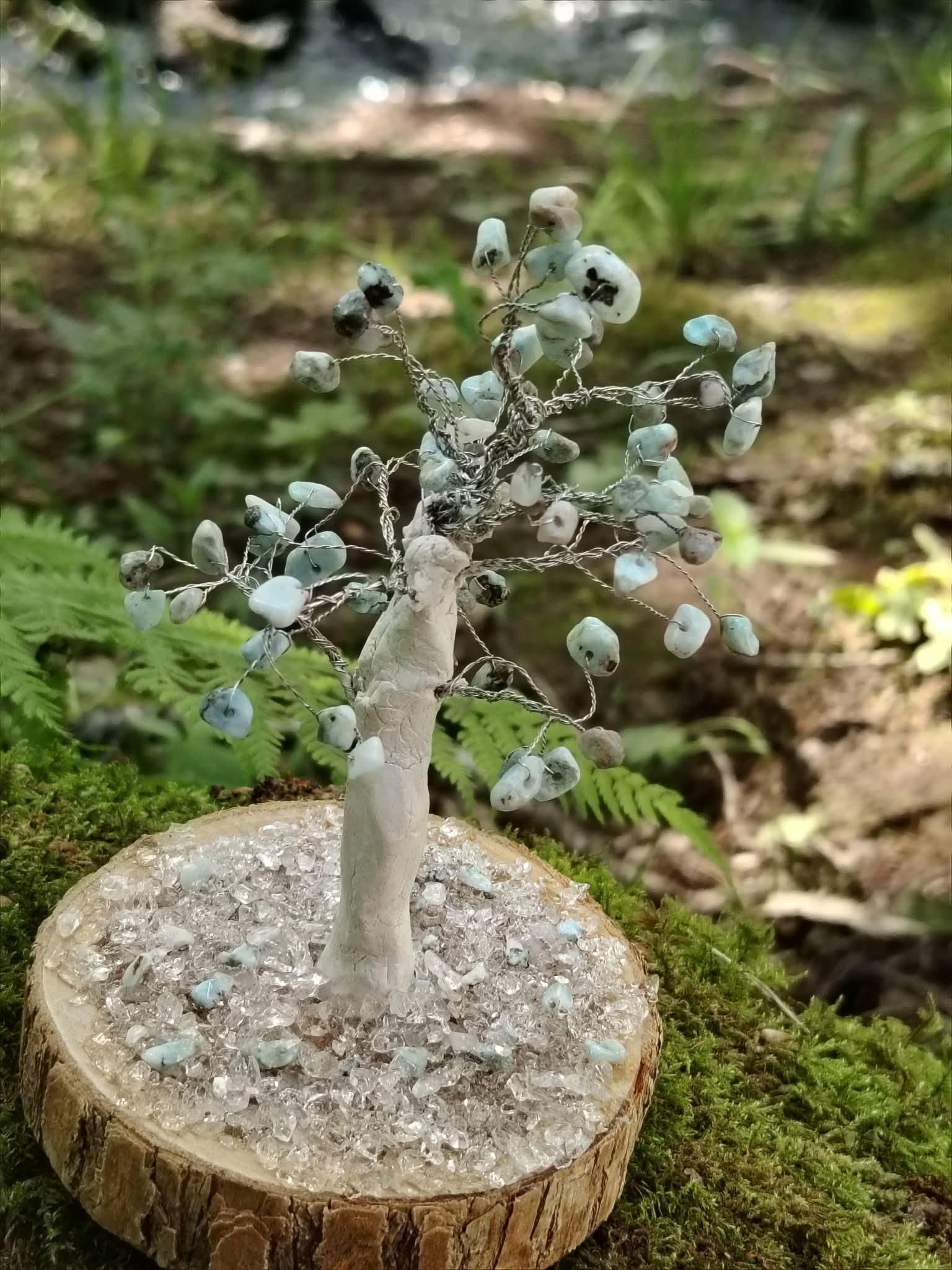 Création Magic Gaïa arbre en larimar sur rondin, pierres bleu pâle et cristal, présenté dans un décor de mousse et ruisseau.