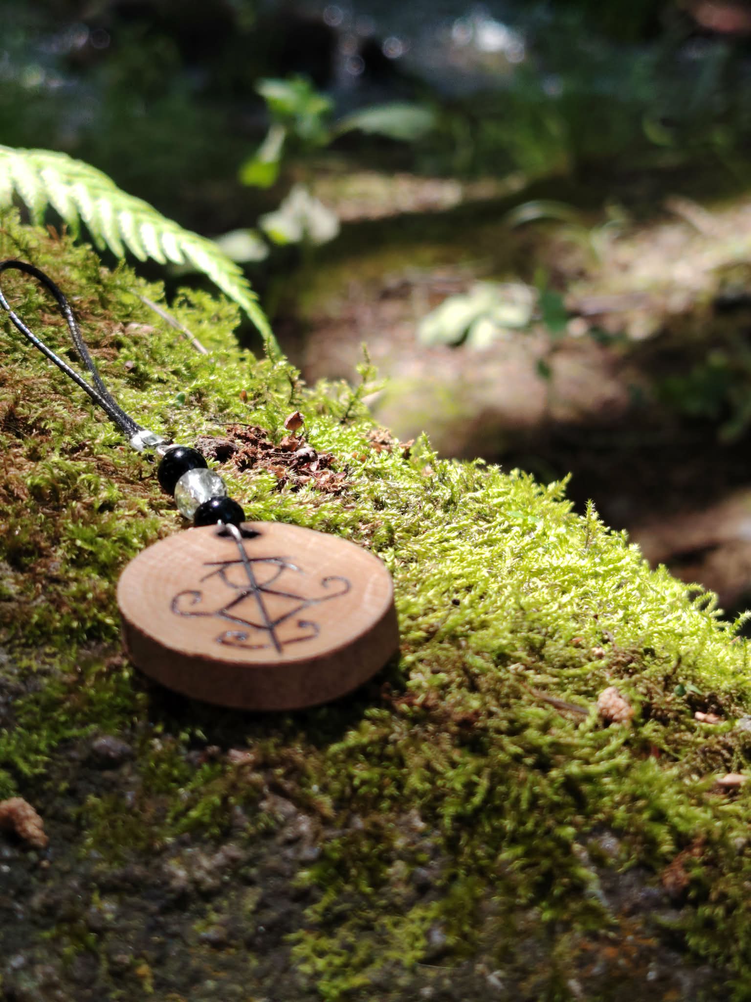 Accessoire Magic Gaïa en bois pyrogravé avec sigil protecteur, tourmaline noire et cristal de roche parmi la mousse.