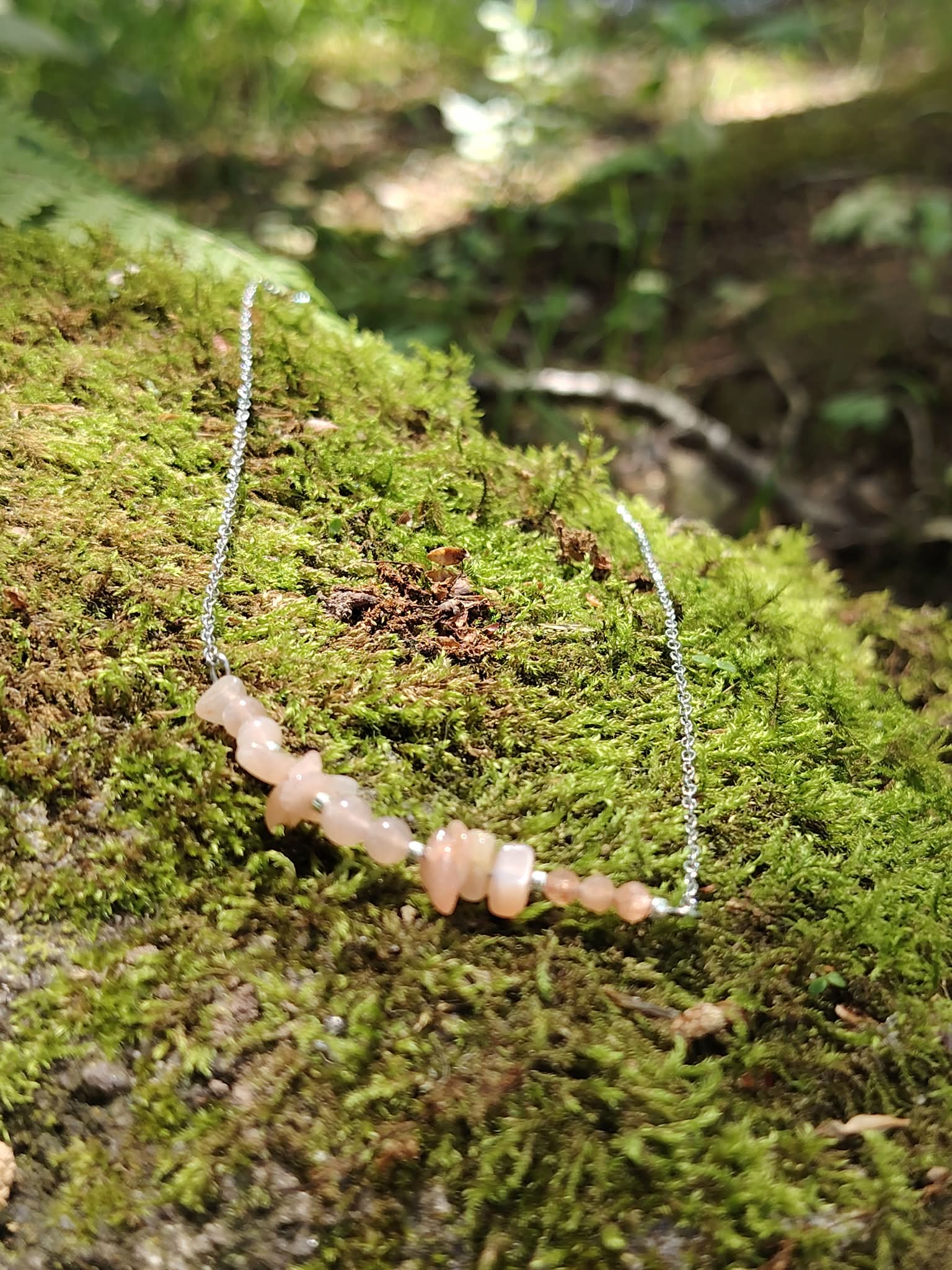 Collier pierre de soleil création Magic Gaïa posé sur mousse en forêt, pierres pêche rosées et chaîne acier inoxydable.