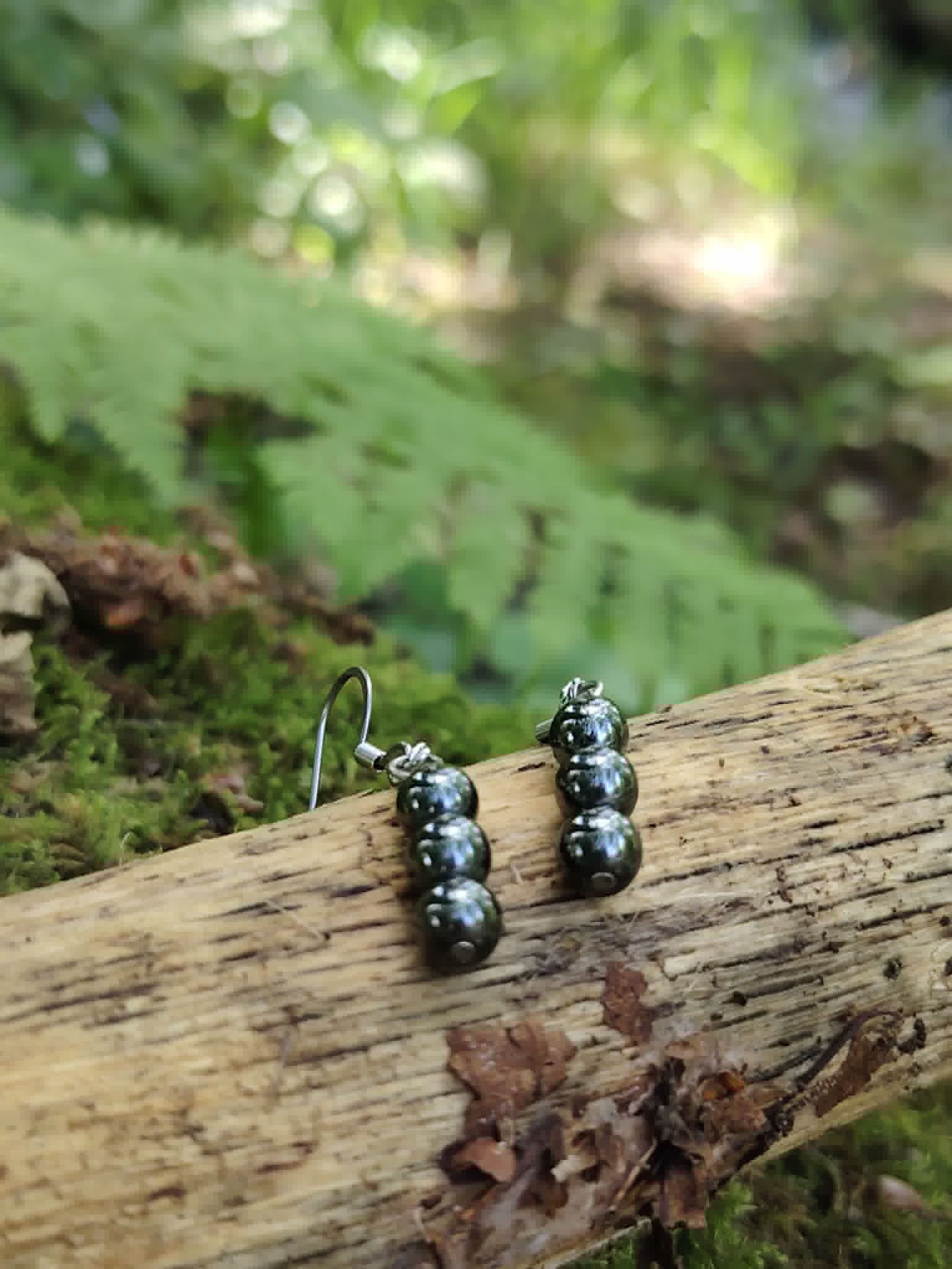 Boucles d’oreilles hématite création Magic Gaïa posées sur branche, mousse et fougère en décor naturel, stabilité intérieure.