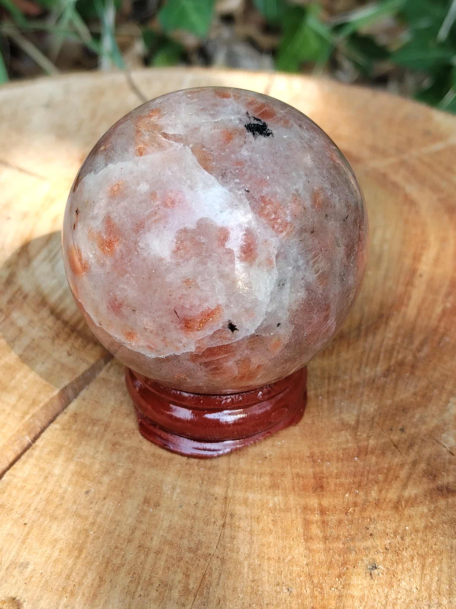 Sphère en Pierre de Soleil naturelle (héliolite) sur support bois, posée sur grand rondin, reflets nacrés orangés, fond végétal flou — symbole de joie, d’optimisme et d’énergie positive.