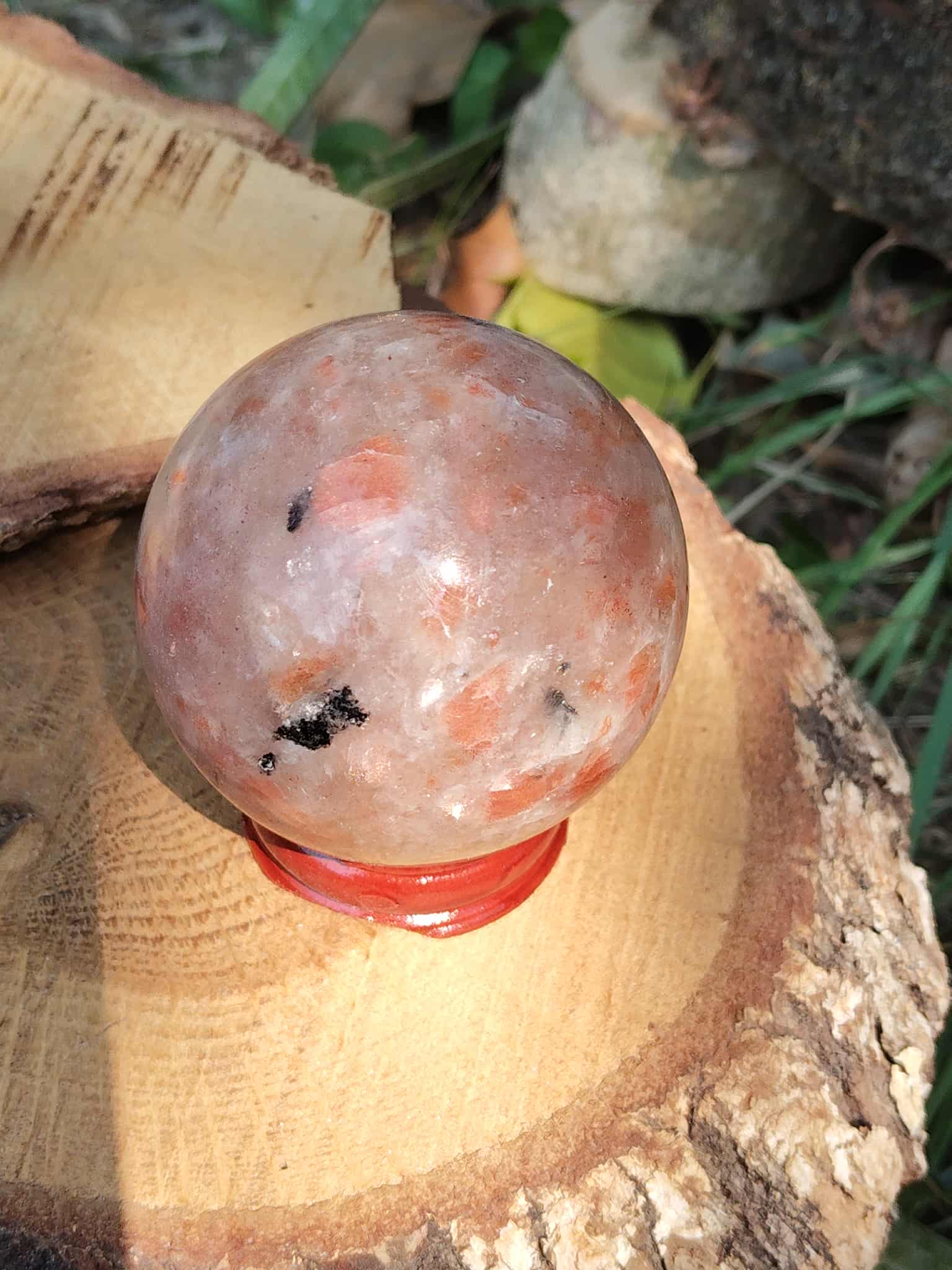 Sphère en Pierre de Soleil naturelle (héliolite) sur socle bois et rondin, reflets nacrés orangés, fond végétal — symbole de joie, d’optimisme et d’énergie positive.