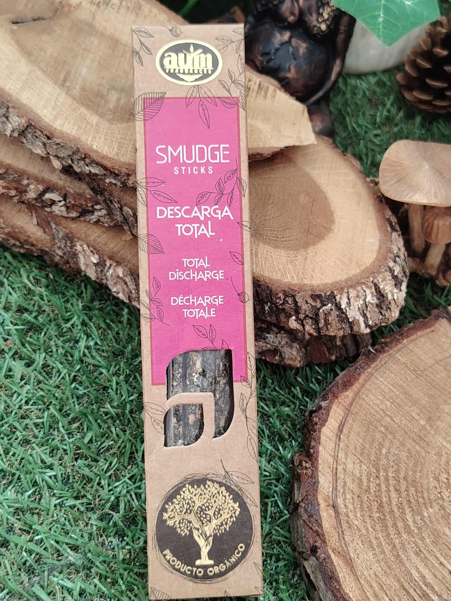 Boîte d’encens Smudge Sticks « Descarga Total » (marque AUM) posée sur rondins de bois et pelouse — encens organique pour rituel de purification/nettoyage énergétique.