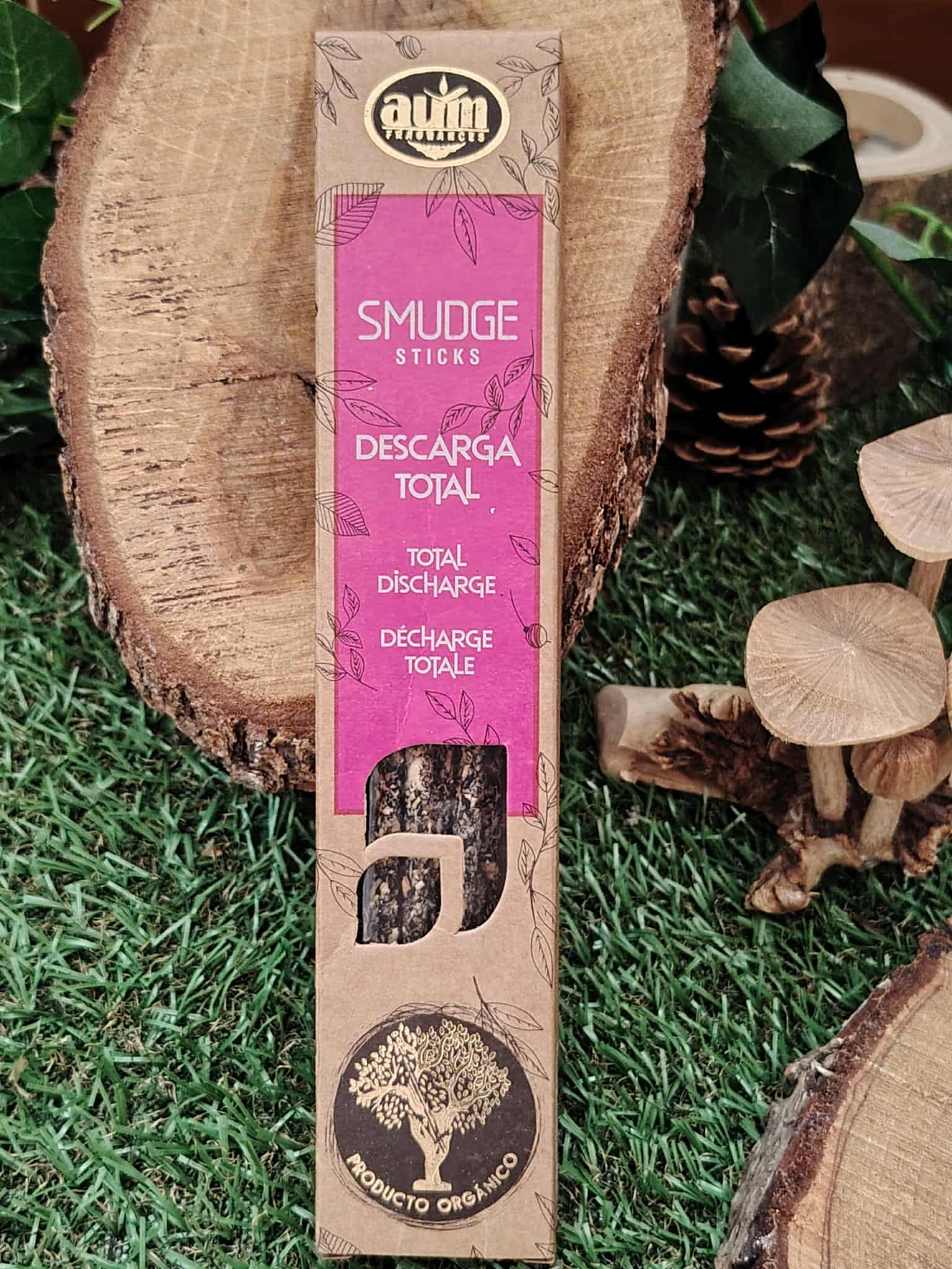 Boîte d’encens organique Smudge Sticks « Descarga Total » (marque Aum) — pack rose/beige posé sur rondins de bois, fond gazon — encens pour rituel de purification