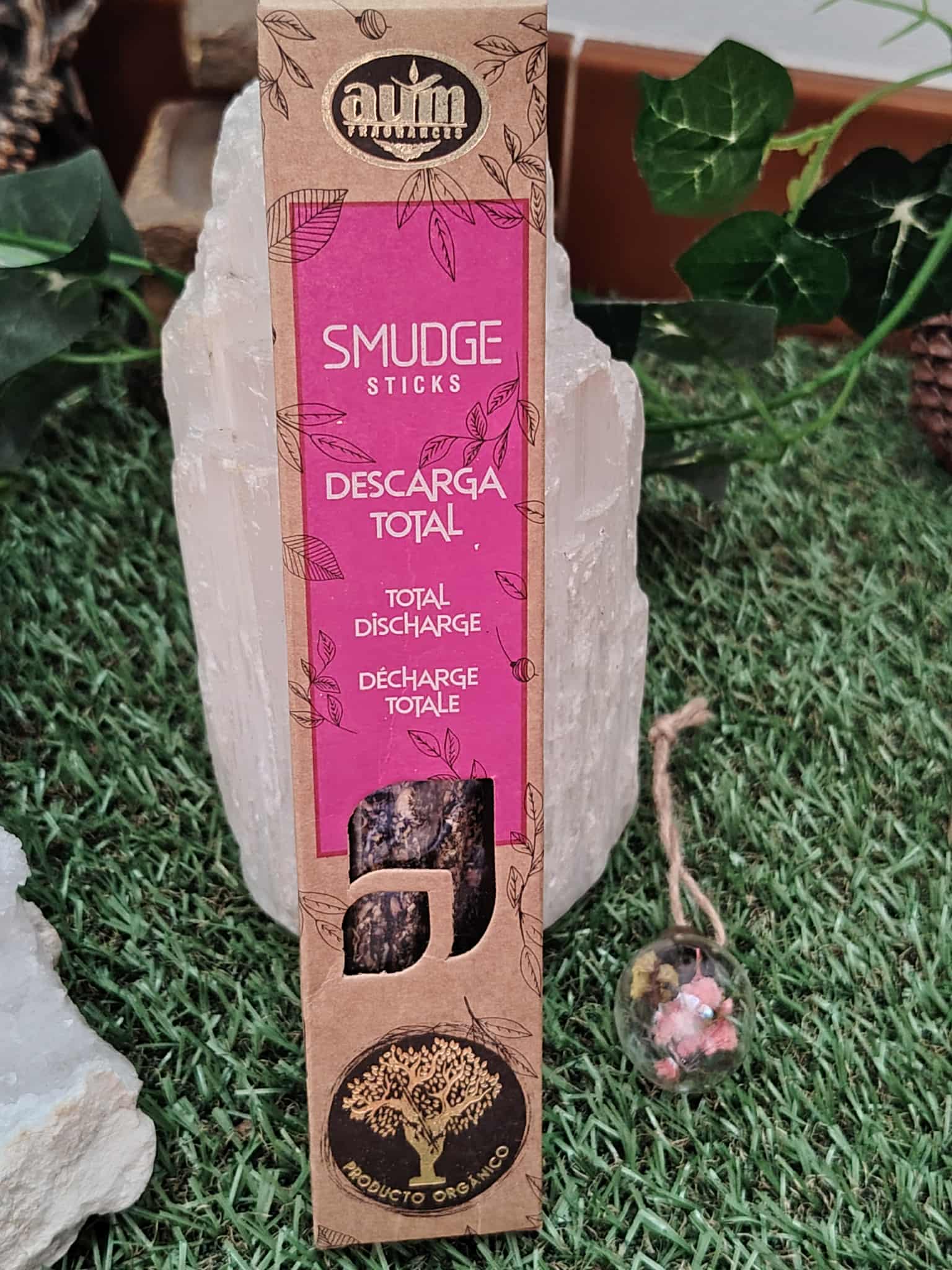 Boîte d’encens organique « Descarga Total / Total Discharge » (smudge sticks) pour rituel de purification, posée sur gazon, adossée à une sélénite, avec une petite décoration rose à côté — packaging rose/beige, fond naturel.