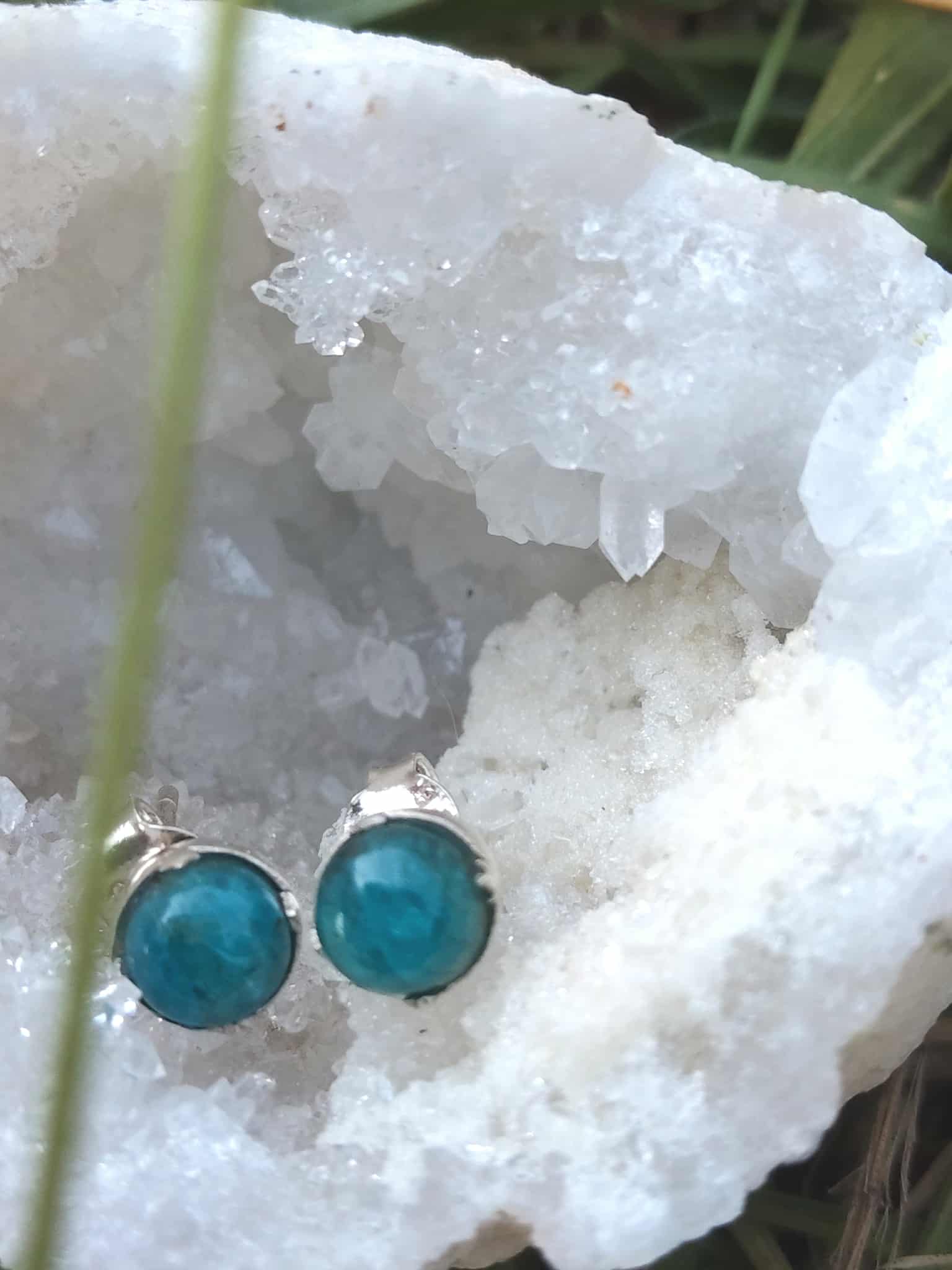 Boucles d’oreilles apatite bleue cabochons ronds en argent 925, posées sur druse de quartz blanc en extérieur (brin d’herbe au premier plan) — clarifie les idées et renforce l’expression créative.