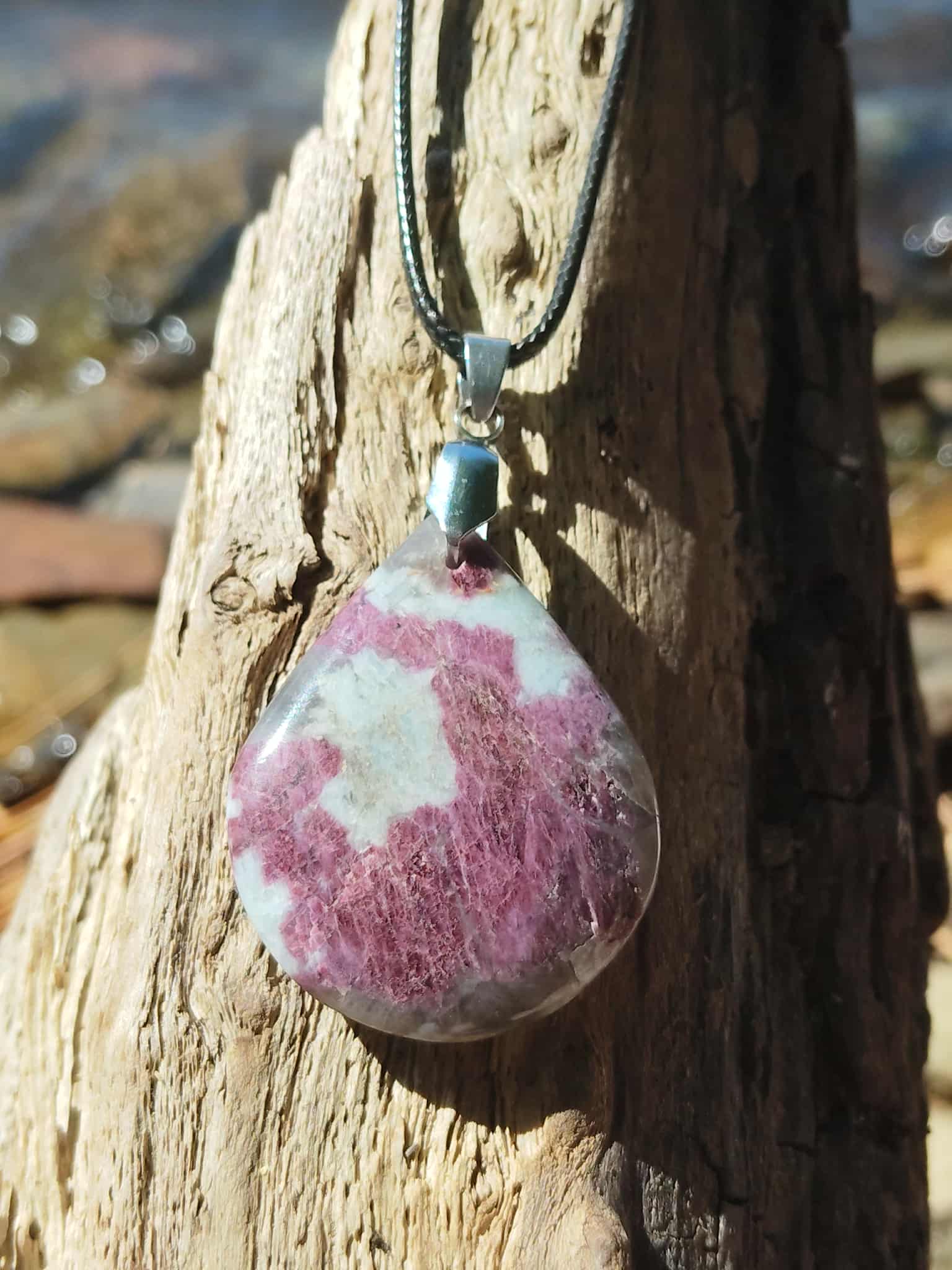 Pendentif en tourmaline rose naturelle, forme ronde polie sur cordon tressé, posé sur bois flotté en extérieur, pierre naturelle associée à la tendresse, au réconfort et à l’équilibre émotionnel.