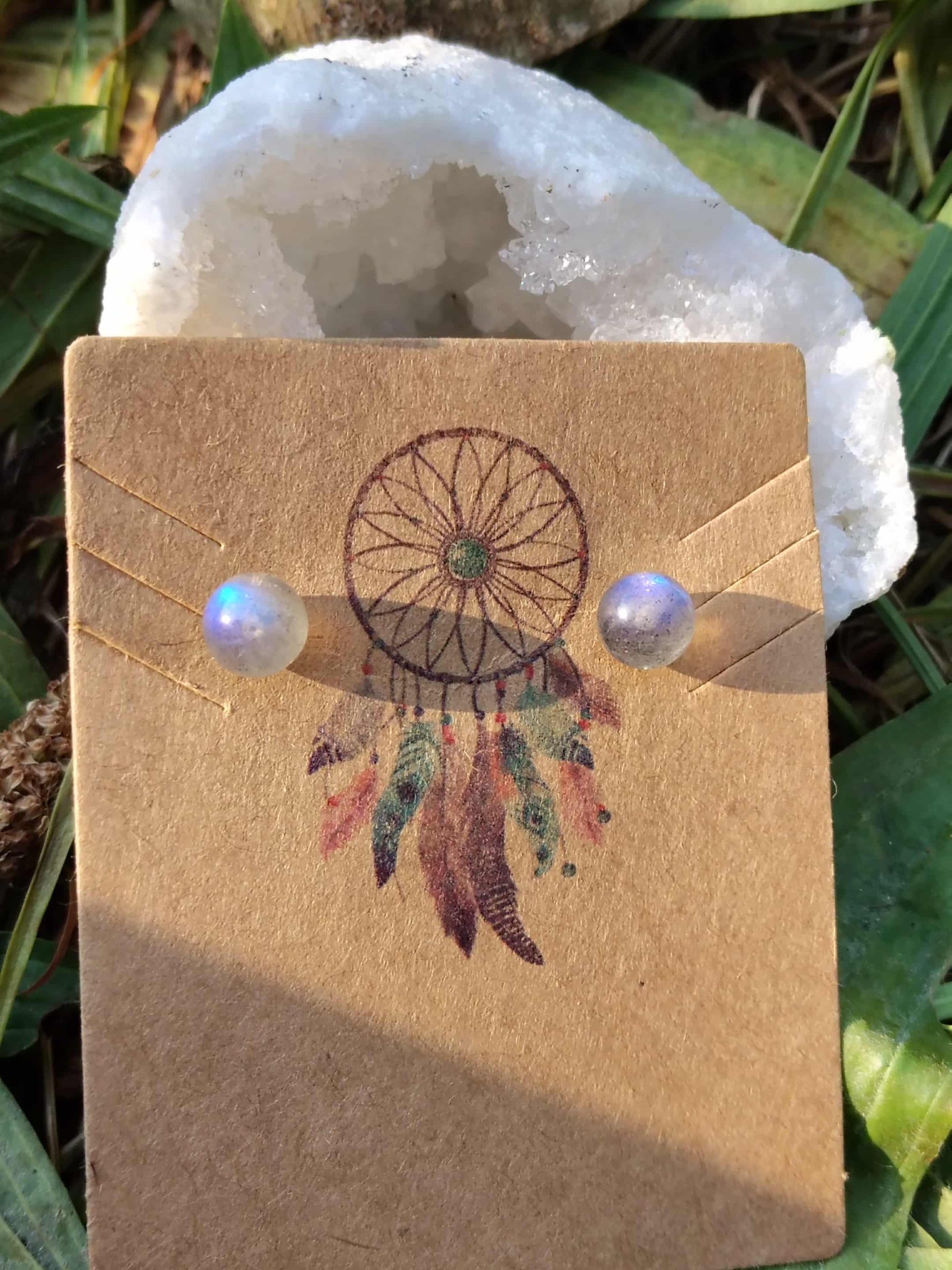 Puces d’oreilles en labradorite bleue, présentés sur un support kraft à motif attrape-rêves devant une géode blanche ; pierre d’intuition, de protection énergétique et d’harmonie intérieure.