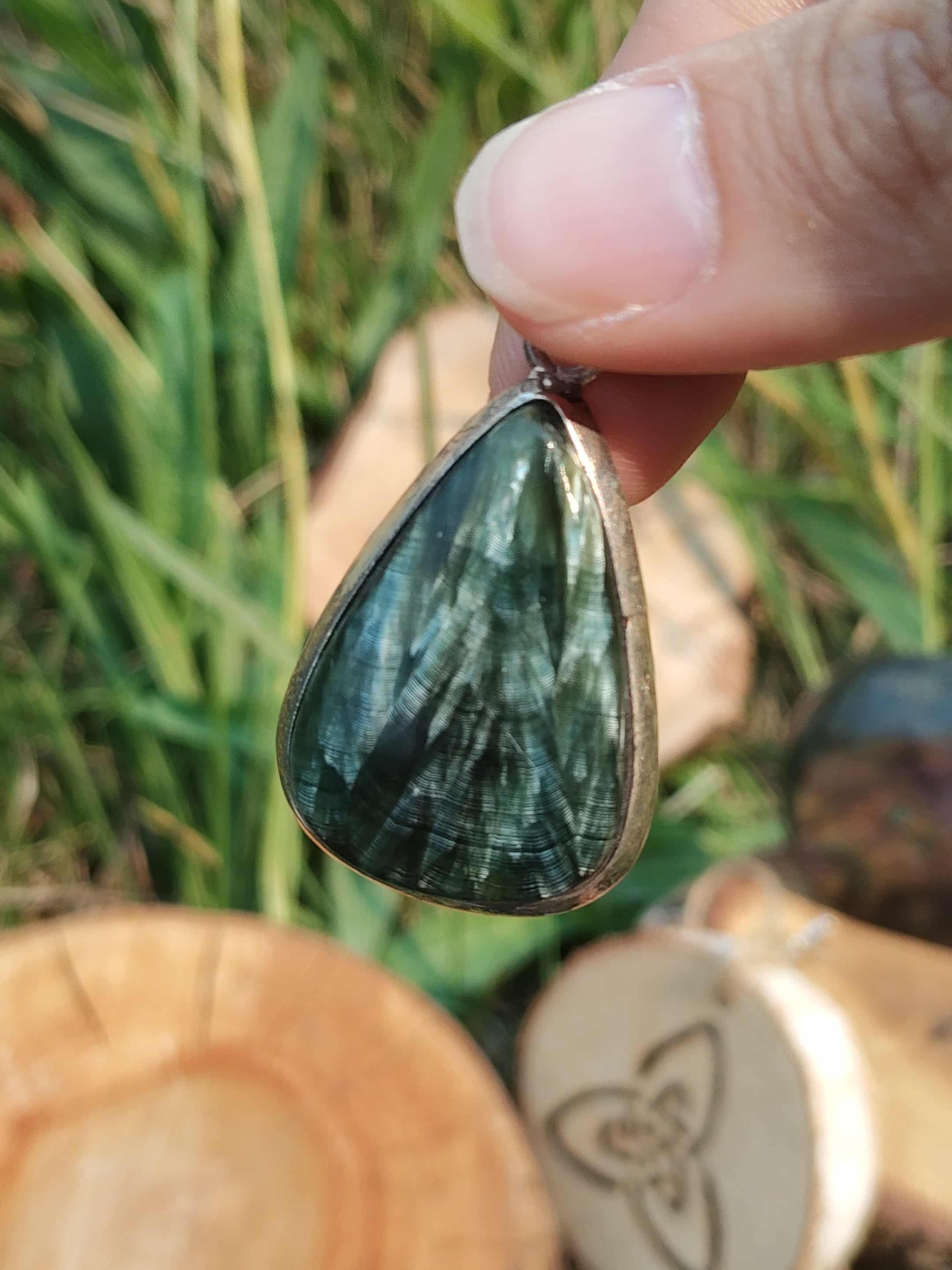 Bijou en séraphinite véritable cabochon goutte en argent 925, présenté en main devant herbes et rondins, pierre naturelle liée au recentrage, à l’écoute de soi et à l’harmonie intérieure.