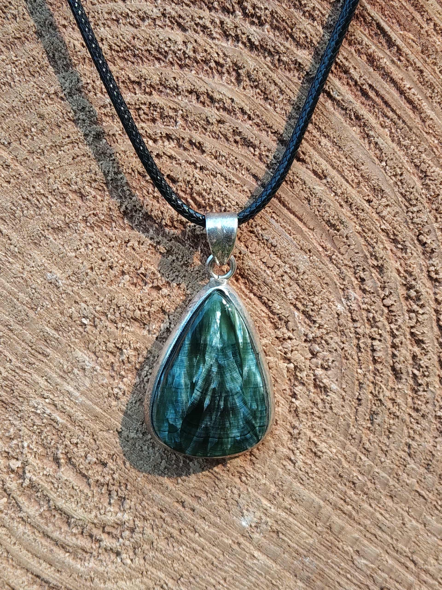 Pendentif en séraphinite véritable à texture fibreuse avec bélière argent, posé sur bois brut en extérieur, bijou en pierre naturelle évoquant la douceur, la paix émotionnelle et la présence à soi.