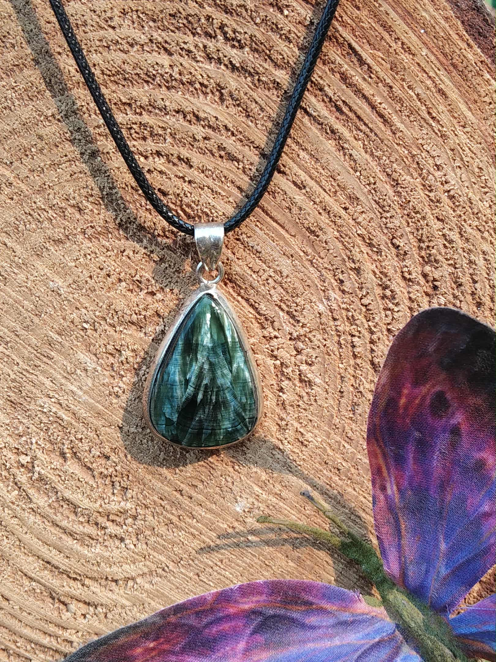 Pendentif goutte en séraphinite posé sur tranche de bois avec décor violet, reflet fibreux et monture argent 925, pierre naturelle associée à la sérénité, au relâchement et à l’équilibre intérieur.