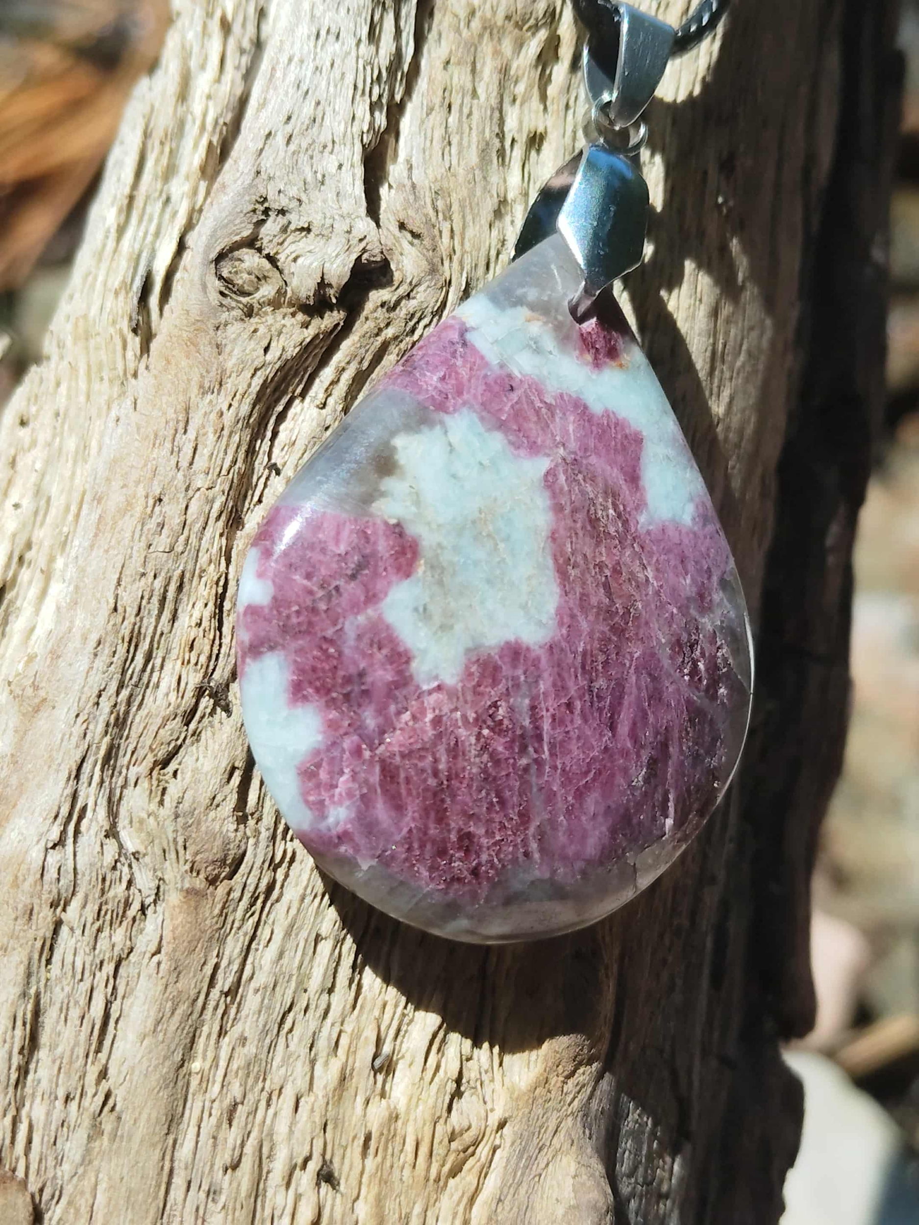 Pendentif tourmaline rose naturelle en goutte avec bélière en acier inoxydable, posé sur tronc de bois avec cordon noir visible, bijou en pierre naturelle lié à la tendresse, au calme affectif et à l’harmonie intérieure.