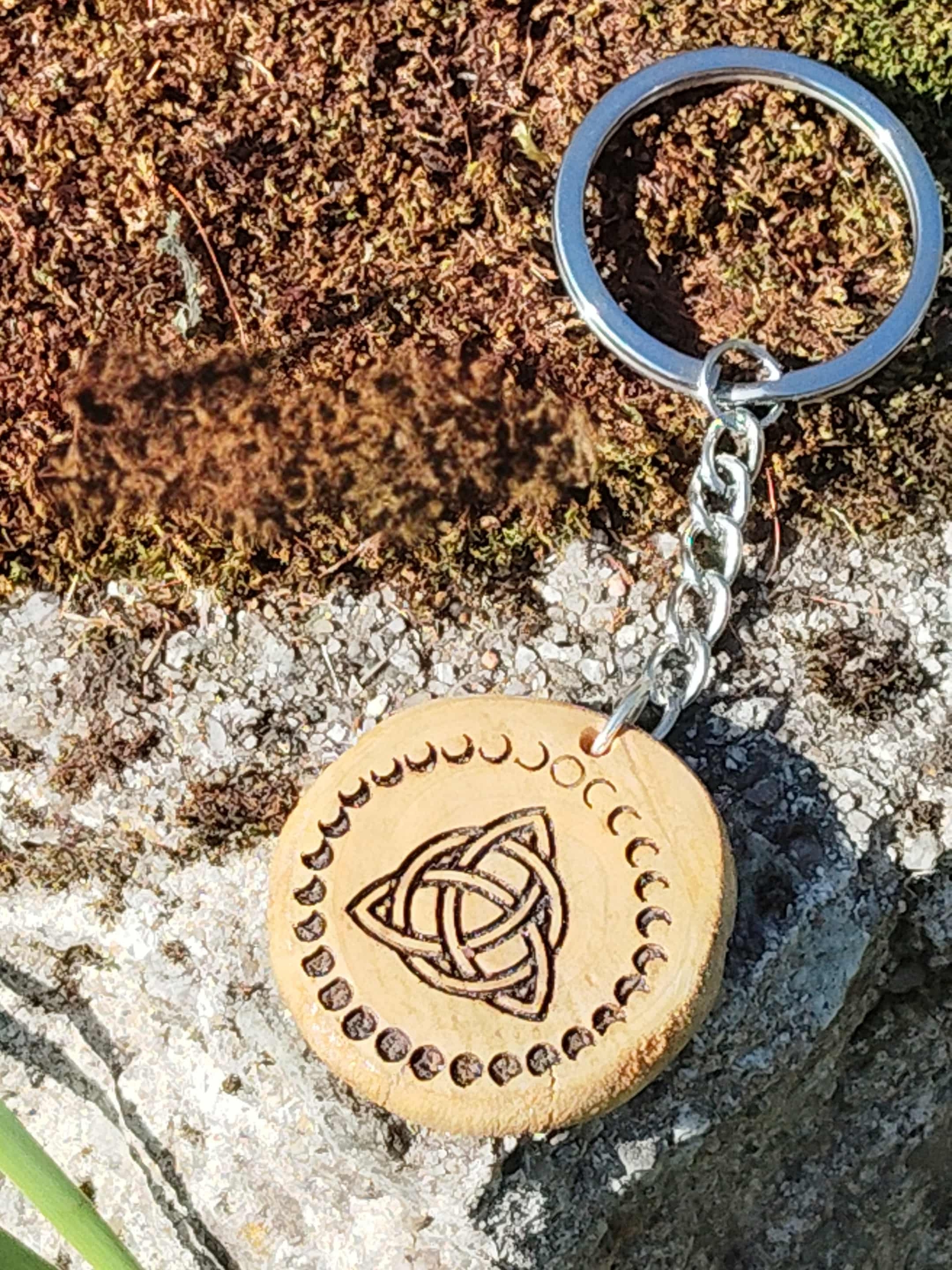 Porte-clés en bois rond gravé triquetra celtique et phases de lune, posé sur mousse et rocher