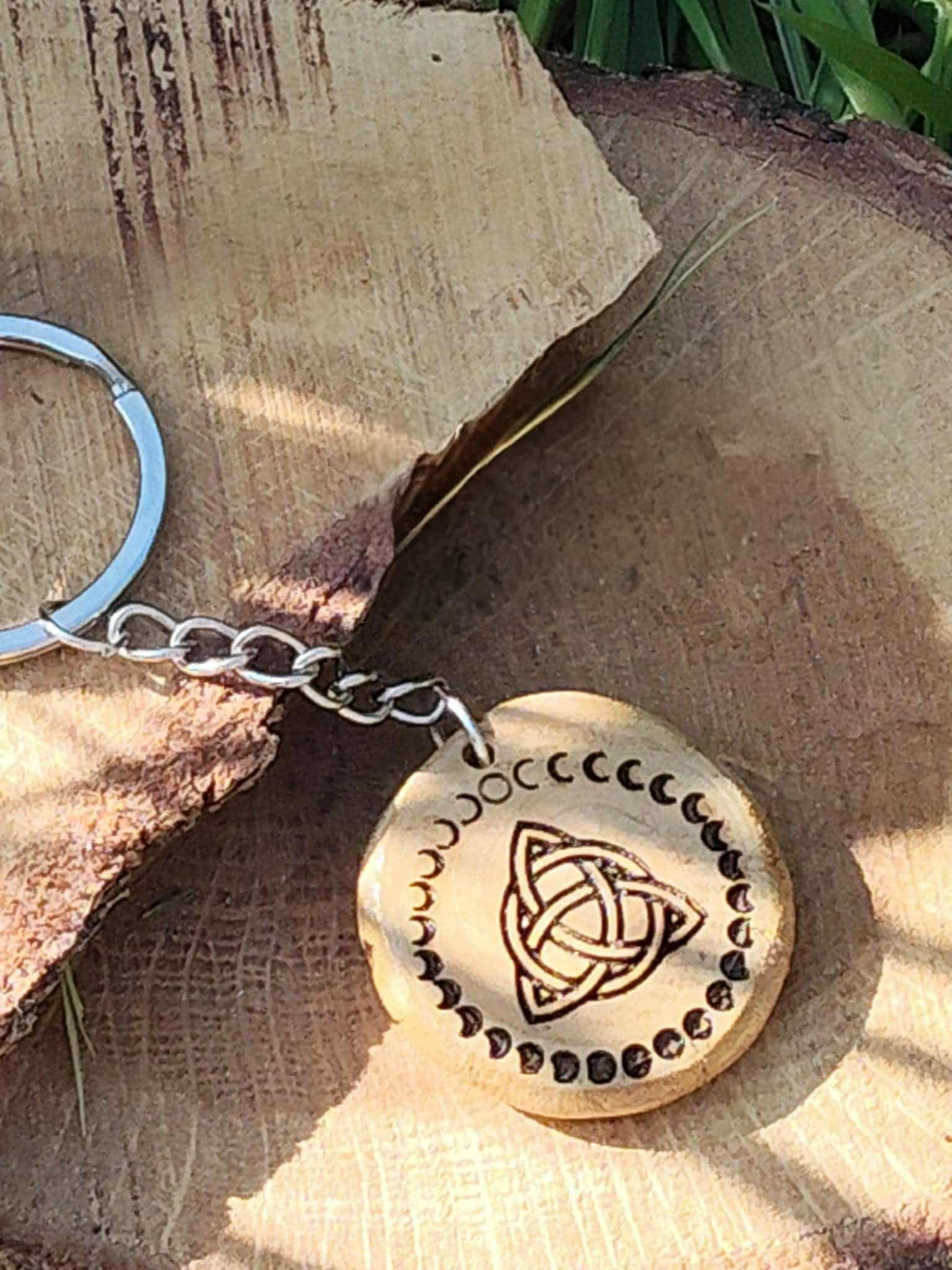 Porte-clés en bois rond gravé triquetra et phases de lune, posé sur rondin avec écorce et brins d’herbe.