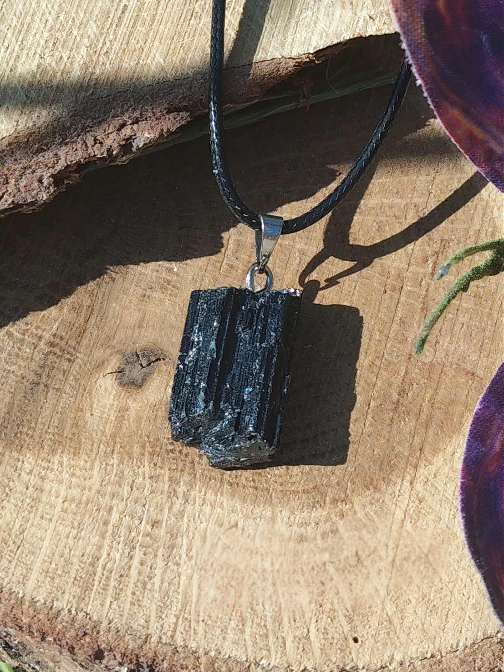 Pendentif tourmaline noire brute sur rondin avec cordon noir et feuilles violettes en arrière-plan, pierre naturelle associée à la stabilité, au recentrage et à la protection énergétique.