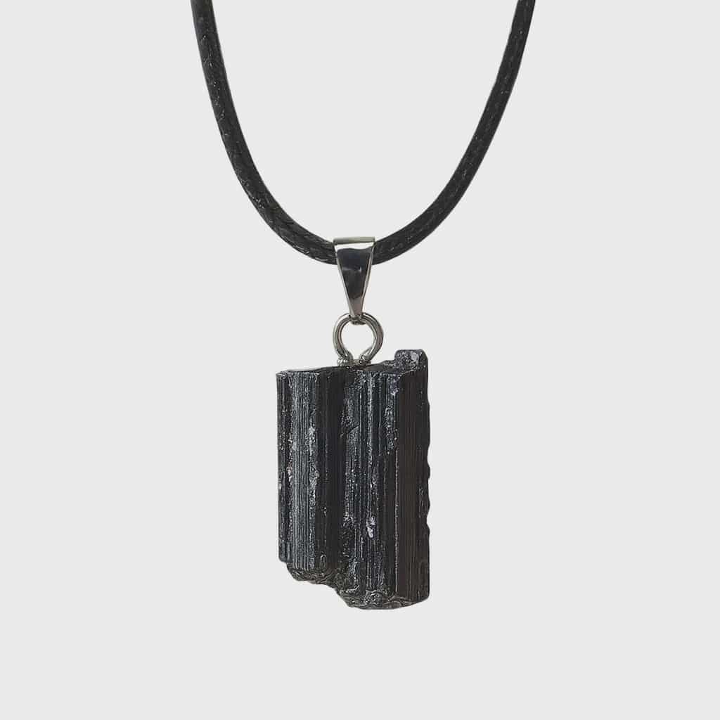 Pendentif tourmaline noire brute sur fond blanc avec cordon noir et bélière acier inoxydable, pierre naturelle reliée au recentrage, à la force calme et à l’équilibre intérieur.