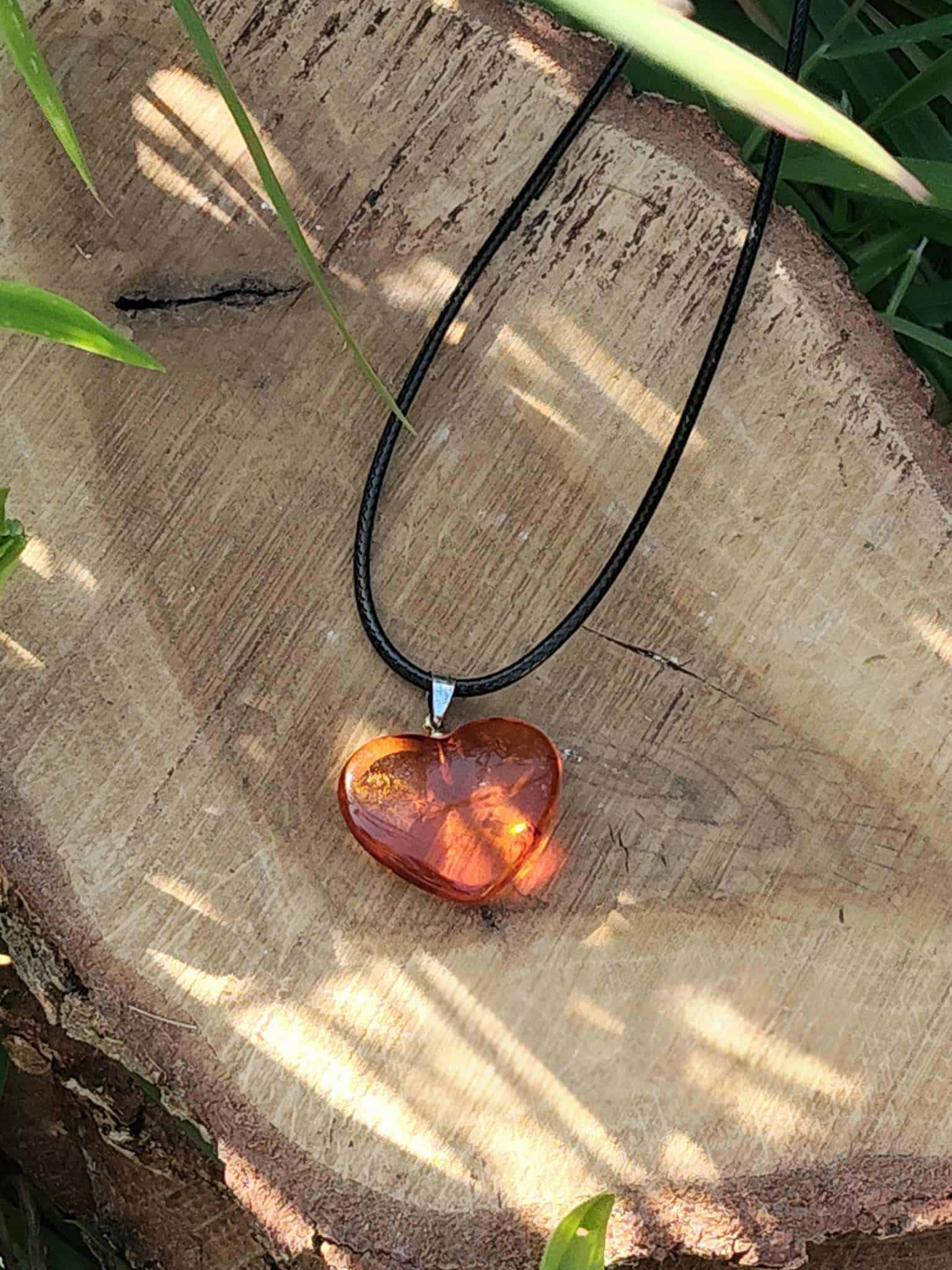 Pendentif cœur en ambre naturel sur cordon noir, posé sur une tranche de bois au soleil, bijou lumineux relié au réconfort, à la sérénité et à l’énergie positive.